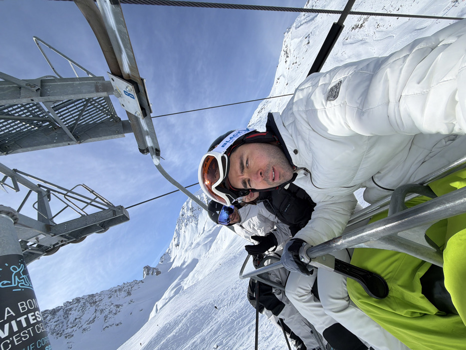 Three individuals are depicted in a selfie-style photograph taken from a ski lift ascending a snow-covered mountain in Courchevel, France, under a clear blue sky. The person in the foreground, a male with dark hair, wears a white padded ski jacket, black ski helmet with an orange-tinted goggles pushed onto it, and bright green ski pants. He holds the ski lift's safety bar with his right hand and looks directly at the camera. Behind him, a second individual in a white ski jacket and blue-tinted goggles raises a gloved hand. A third, partially obscured individual in a dark jacket is visible behind the second. Overhead, the metal structure of the ski lift tower and cables, including a component marked with the number "8", are prominent. Steep, snow-covered slopes extend into the background.