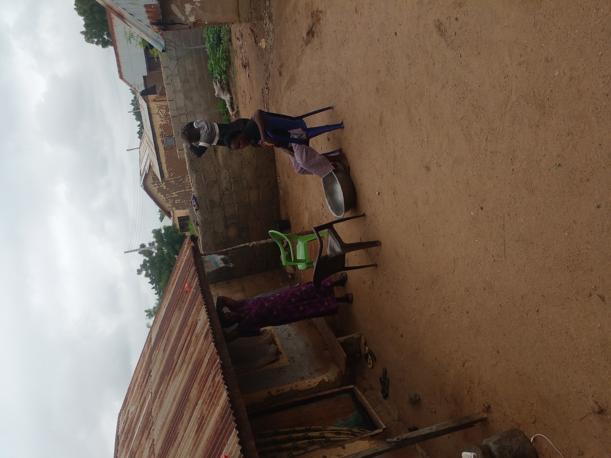 In the city of Unknown, Unknown, an outdoor scene unfolds under a cloudy sky. A woman in a long, purple patterned dress stands near the entrance of a weathered building with a rusty corrugated metal roof. She has her hands behind her head, possibly scratching her scalp. In front of her, a dark plastic chair is positioned near the building's entrance, and a bright green plastic chair is slightly to the right. Further to the right, a young woman is seated on a blue plastic chair, wearing a black t-shirt and a patterned skirt. In front of her, a large metal basin sits on the sandy ground. To the right of the seated woman, a wall constructed of cinder blocks encloses an area with some greenery and what appears to be a piece of driftwood. In the background, other buildings with similar corrugated metal roofs and mud-plastered walls are visible, suggesting a rural or semi-urban setting. The ground is covered in reddish-brown sand.