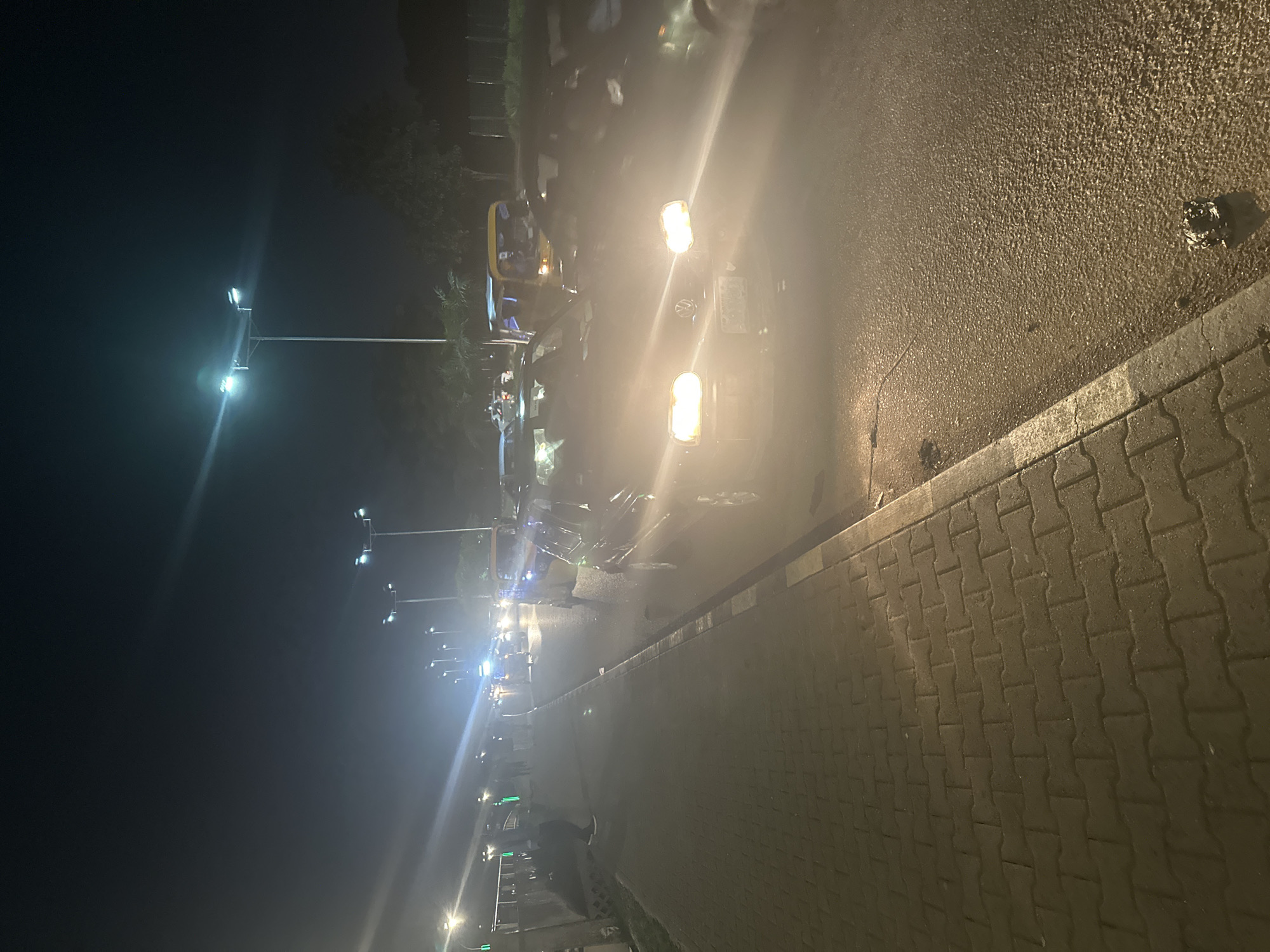 This image depicts a nighttime scene on a city street, likely in Abuja, Nigeria, characterized by artificial lighting from street lamps and vehicle headlights. The perspective is at street level, looking down a road with a paved sidewalk to the left. Several vehicles are visible, some with their headlights on, suggesting traffic or a parked vehicle. The road surface appears wet or reflective, possibly due to recent rain or dampness, contributing to the diffused lighting.
In the foreground, the paved sidewalk is made of interlocking concrete blocks, with a curb separating it from the road. To the right of the sidewalk, the asphalt road is visible, with some debris or trash present on the surface.
Further down the road, a line of vehicles is suggested by the illuminated headlights. A yellow auto-rickshaw, commonly known as a "Keke Napep" in Nigeria, is prominent among the vehicles. A dark-colored car with its headlights on is in the middle ground, and other vehicles are visible in the background, their forms softened by the low light and atmospheric conditions.
Several figures of people are discernible on the sidewalk and near the vehicles, their presence indicating activity on the street. They appear to be walking or standing, their figures partially obscured by the lighting and distance. Streetlights, tall poles with multiple lamps, are spaced along the road, providing illumination and contributing to the overall nighttime ambiance. The sky is dark, typical of nighttime, with some lens flare effects from the bright lights. The environment appears to be an urban or semi-urban setting, with the presence of infrastructure like roads, sidewalks, and streetlights.