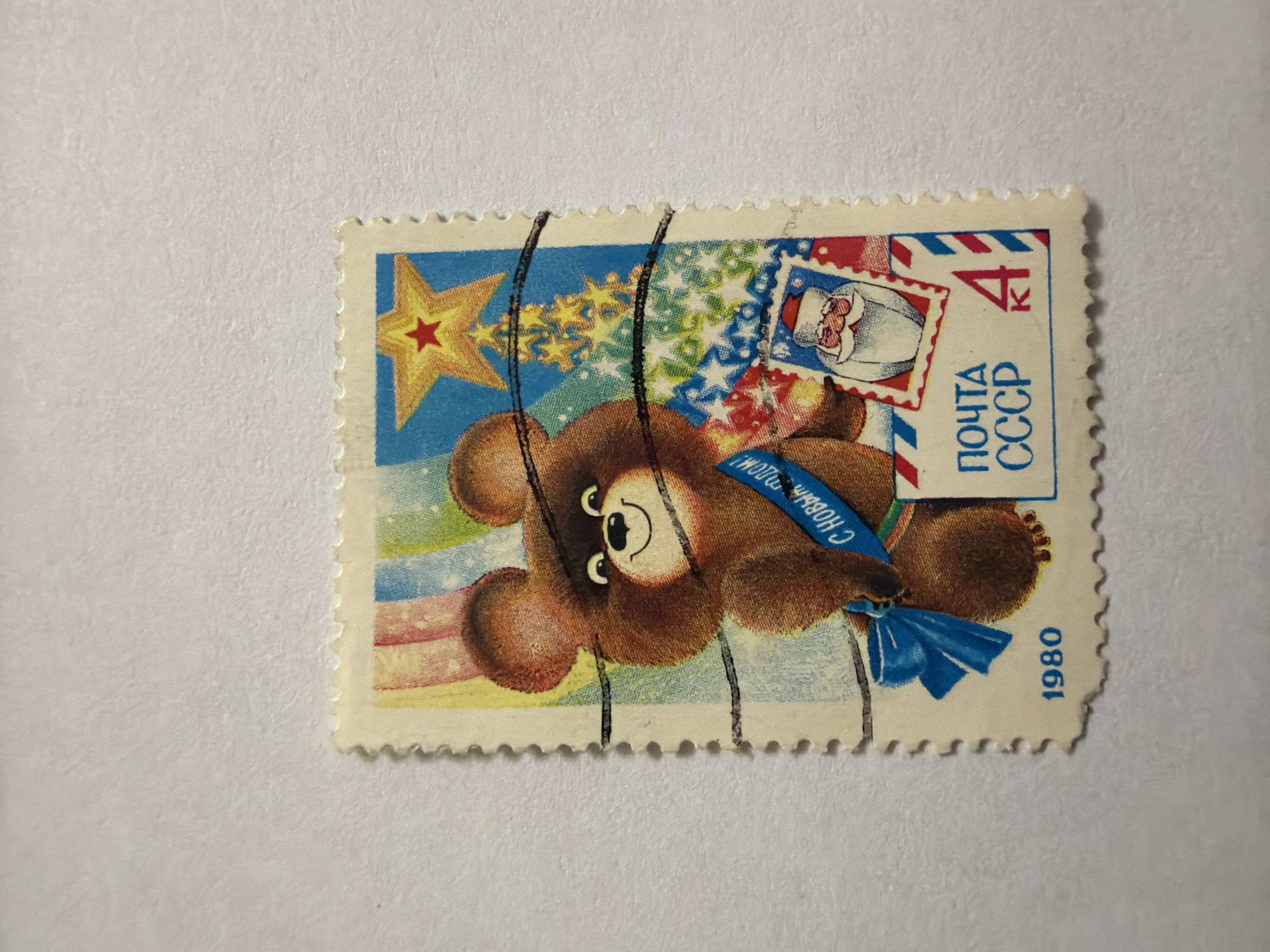 The image shows a Soviet Union (USSR) postage stamp issued in 1980. It features an illustration of the Olympic Mishka bear, a symbol of the 1980 Moscow Olympics, wearing a blue sash with the text "C Новым Годом!" (Happy New Year!). The stamp also includes a Christmas tree with a red star, an image of Santa Claus, the price "4 K" (4 Kopeks), and the inscription "ПОЧТА СССР" (Postal Service of the USSR). The stamp is torn around the edges, and several black vertical lines cross the image.
Izhevsk, Russia is not related to this stamp.