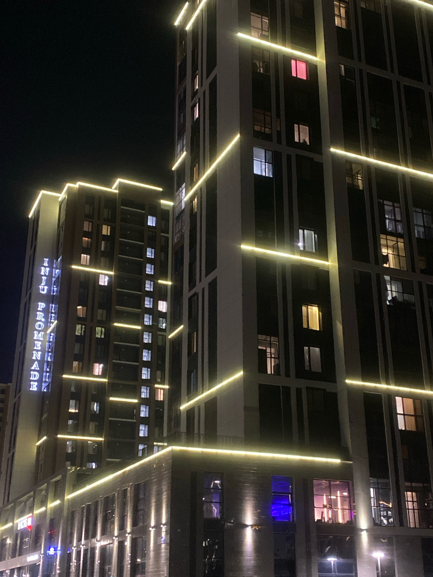 This is a nighttime, vertical, medium shot of the "INJU PROMENADE" building complex in Astana, Kazakhstan. The complex consists of two main buildings, both tall and modern, with dark facades and numerous windows. The buildings are outlined with bright yellow LED strip lighting, which accentuates their architectural lines and creates a striking contrast against the dark night sky.
The left building has the words "INJU PROMENADE" vertically displayed in blue neon letters on its side. The windows of both buildings show varying levels of interior light, indicating occupancy. Some windows glow with warm white light, while others display cooler blue or pink hues, suggesting different interior lighting or activities. The lower floors of the buildings appear to be commercial spaces, with more prominent lighting and some signage that is partially obscured by the angle and lighting. The overall impression is one of a well-lit, modern urban environment at night.