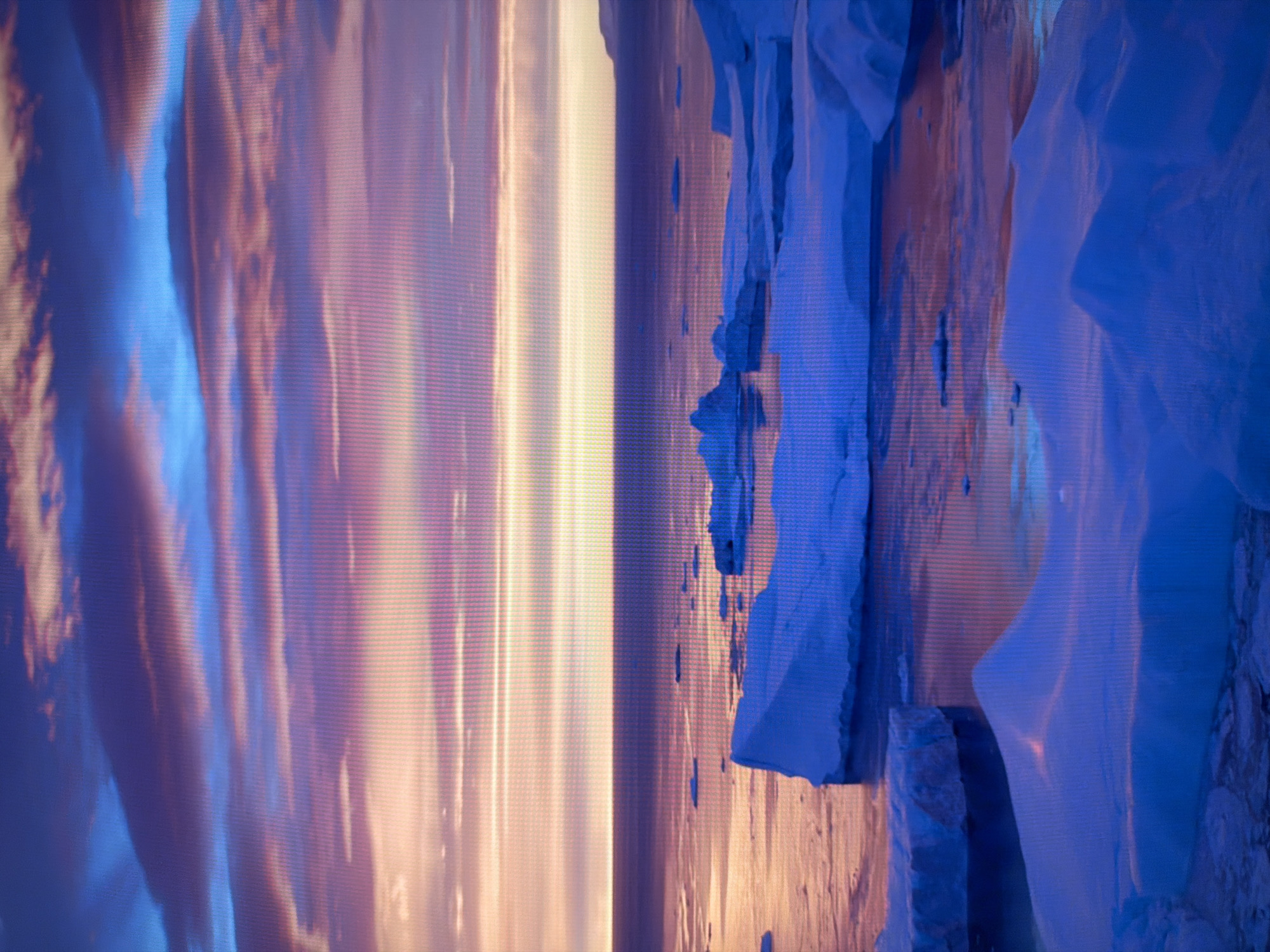 The image displays an arctic landscape at twilight. The sky is filled with dramatic, striated clouds in shades of deep blue, purple, and pale pink, suggesting a sunset or sunrise. Below the sky, a vast expanse of ocean reflects the colors of the sky, with a band of lighter hues indicating the horizon. Numerous icebergs of varying sizes, predominantly white and tinged with blue, are scattered across the water and in the foreground. Some icebergs are large and imposing, while others are smaller and appear to be floating. The water is calm, with reflections of the ice and sky visible on its surface. The overall impression is one of serene, cold beauty. There are no people, buildings, or any indications of urban environments present in this image. The scene is not located in the city of Kyiv, Ukraine.