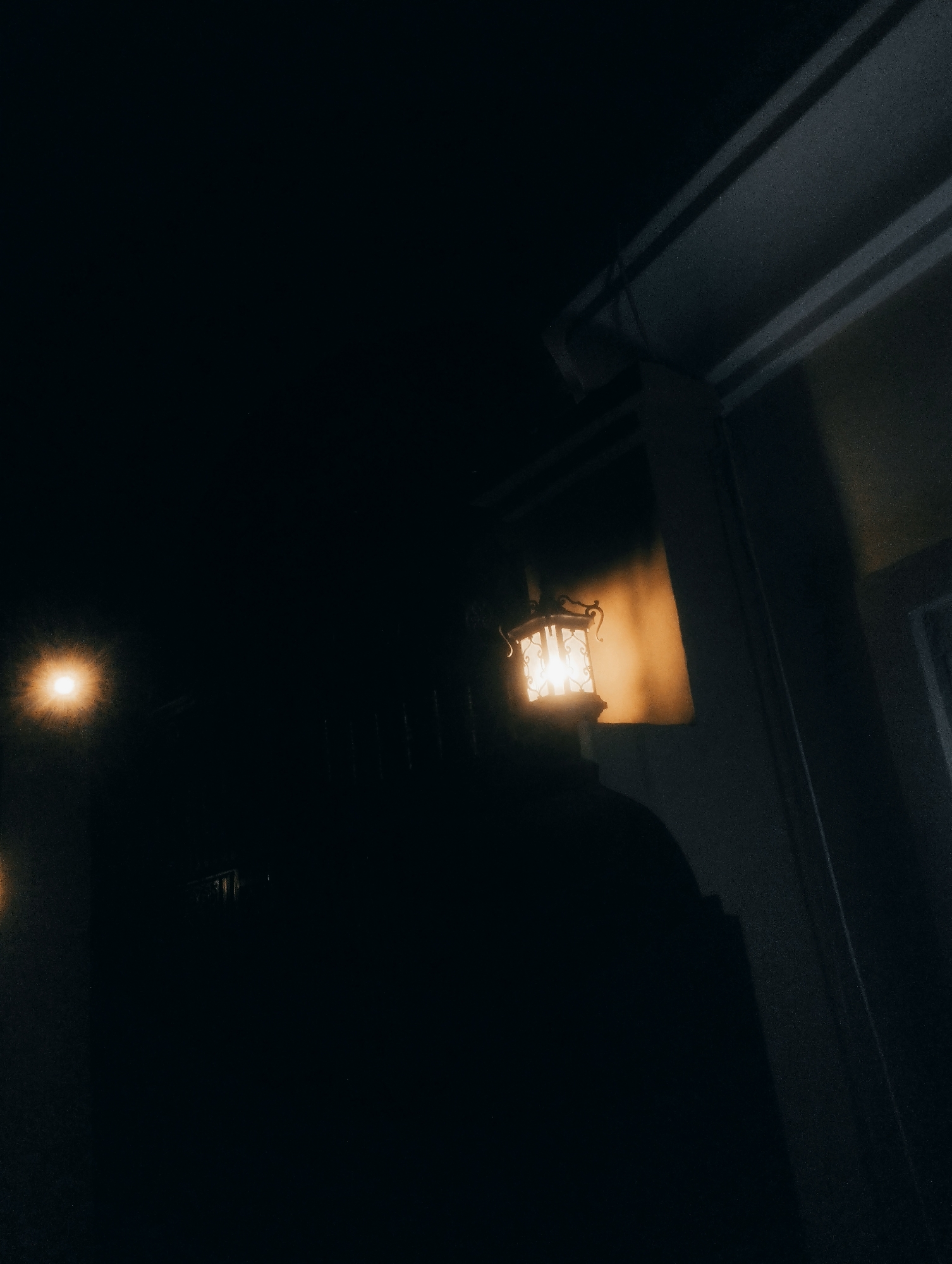 This is a photograph taken at night in Jos, Nigeria, featuring an illuminated outdoor lantern mounted on a building structure. The lantern, with a decorative metal frame and glass panels, emits a warm, orange light. The surrounding environment is predominantly dark, with hints of architectural elements visible, such as a ceiling overhang and pillars. The scene is captured at a low angle, with a significant portion of the image consumed by shadow, suggesting the absence of other significant light sources.