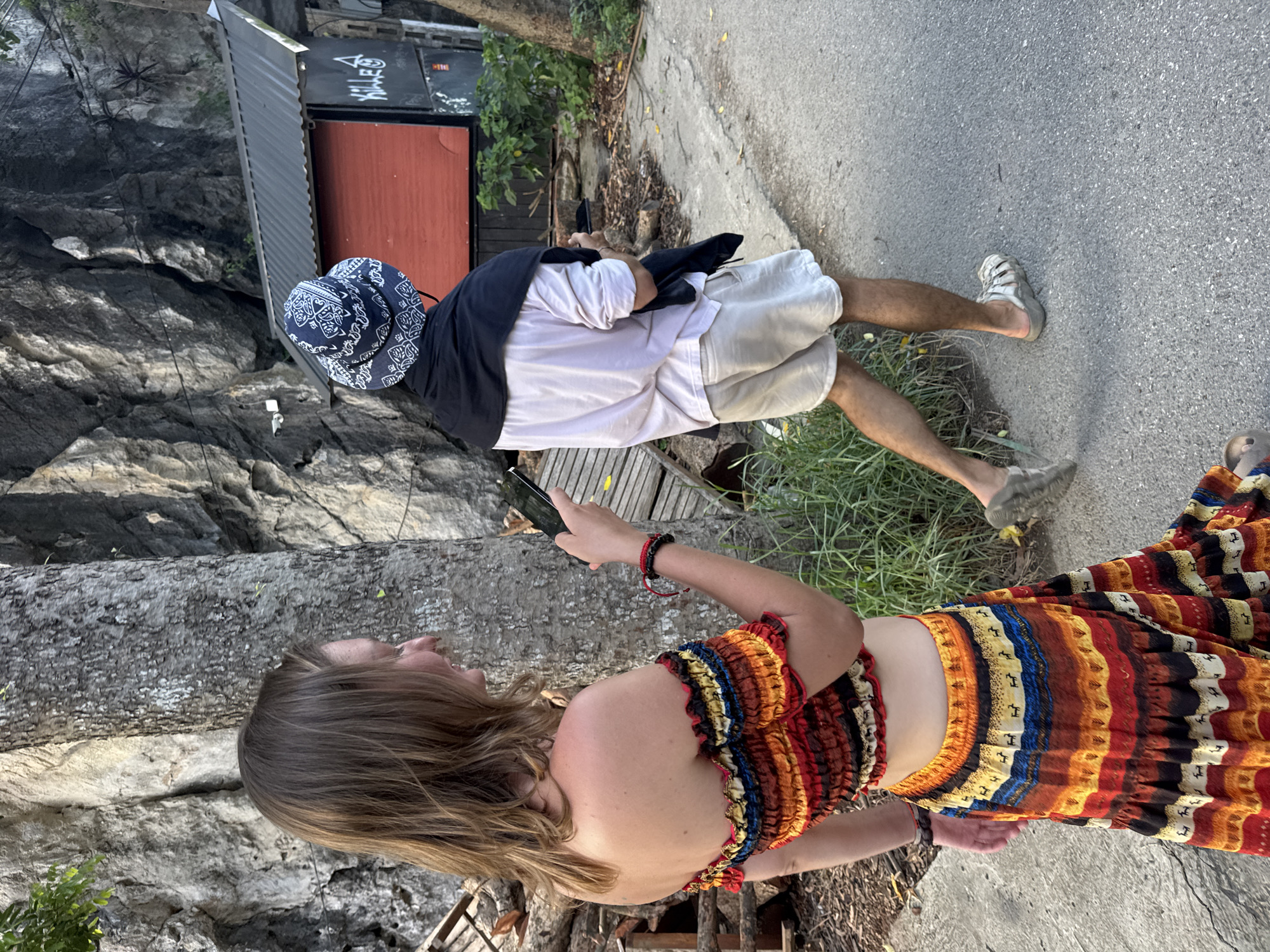 A woman with long brown hair, wearing a colorful off-the-shoulder crop top and a matching tiered skirt with a tribal pattern, is walking away from the camera, holding a smartphone in her right hand. She is on a paved pathway next to a large tree trunk and a rocky cliff face. Behind her, a person wearing a white t-shirt, light-colored shorts, a black jacket draped over their shoulders, and a blue patterned bucket hat is also walking away from the camera. They are holding a smartphone in their left hand. Further in the background, a small building with a dark metal roof and a red facade is visible, with graffiti on its side. The environment appears to be outdoors, possibly in a rural or park-like setting, with vegetation and natural rock formations. The image was taken during the day, with natural light illuminating the scene. The location is identified as Nam Hoa Lư Ward, Vietnam.