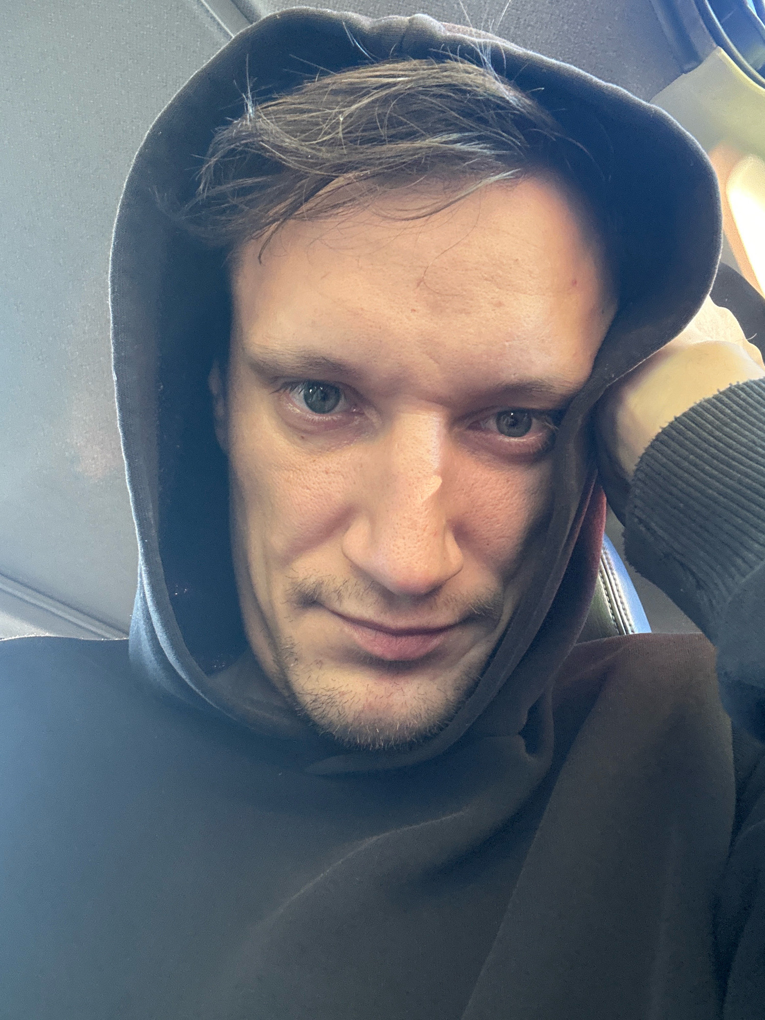 A selfie of a young Caucasian man with brown hair and blue eyes is taken from a low angle, showing him wearing a dark hooded sweatshirt. His left hand is resting on his forehead, and his head is tilted. The background appears to be the interior of a vehicle, possibly a bus or train, with a textured grey headrest visible behind him. The lighting is natural and casts a shadow across his face. The image was taken in Makiv, Ukraine.