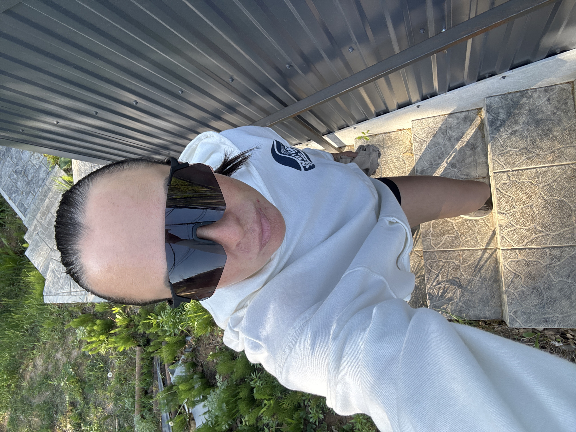 A woman wearing sunglasses and a white hooded sweatshirt stands on gray stone pavers next to a gray corrugated metal wall. The pavers form a small step. Beyond the pavers is green grass and small shrubs. The photograph appears to have been taken outdoors during the day. No specific location in Chaikovsky, Russia, can be determined from the image.