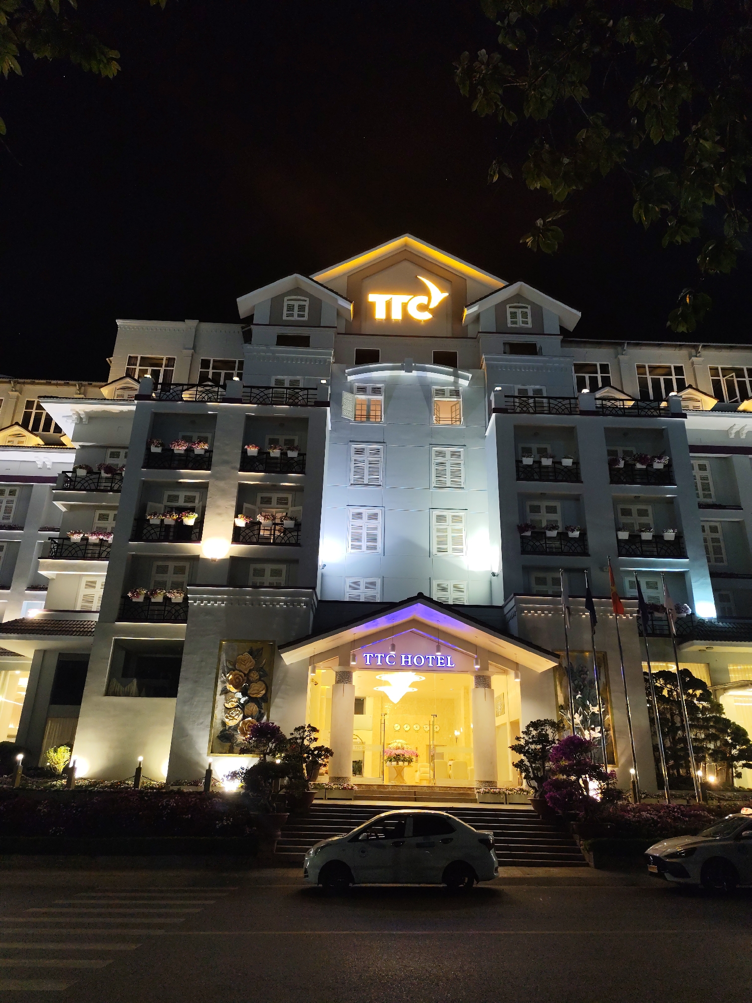 The image shows the TTC Hotel Premium Michelia in Da Lat, Vietnam, at night. The hotel is a multi-story building with a light blue facade, white window shutters, and balconies adorned with flower boxes. The "TTC" logo is prominently displayed in yellow on the roof, with "TTC HOTEL" in purple above the main entrance. A car is parked in front of the hotel, on a crosswalk. Flags stand near the entrance. The building and surrounding landscaping are illuminated by lights. The sky is dark.