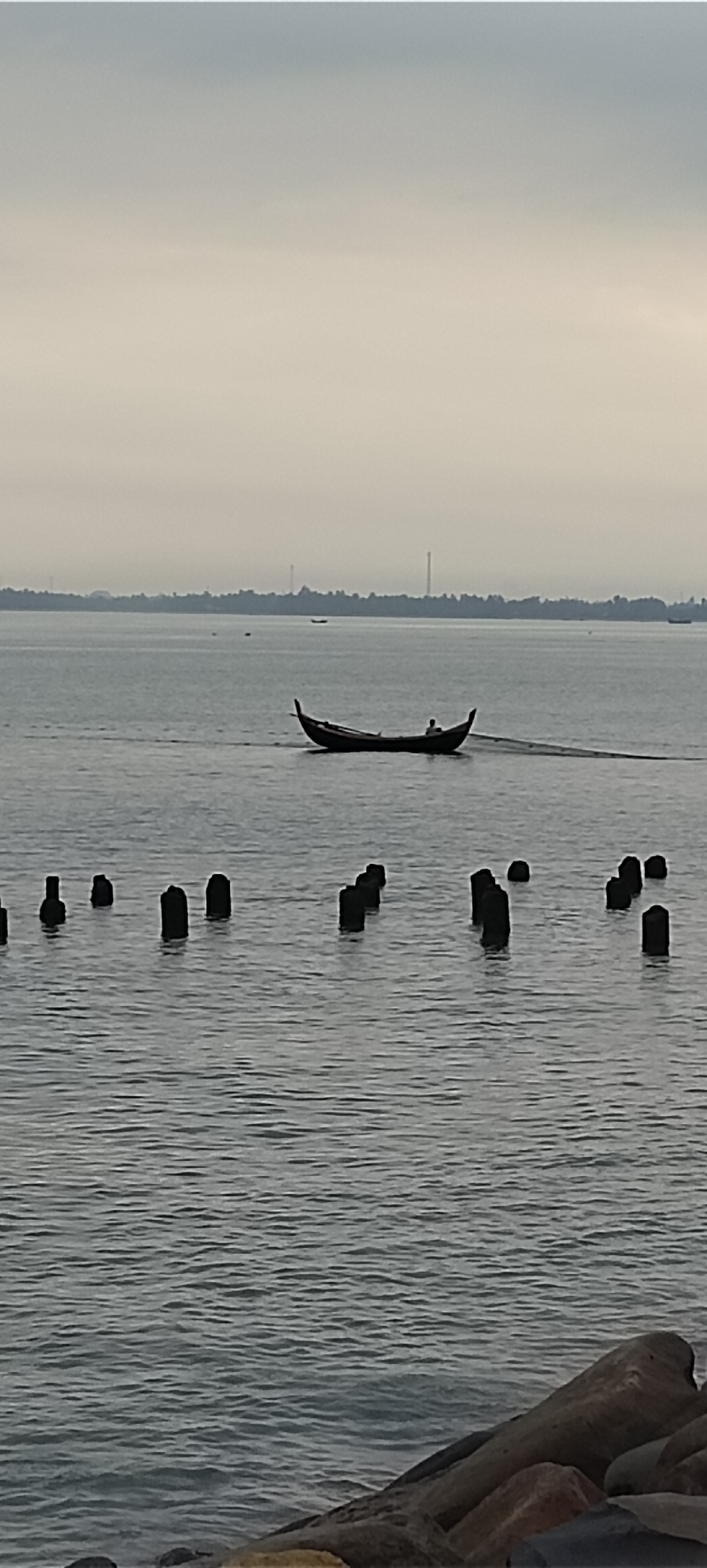 A photograph captured from a slightly elevated position shows a wide expanse of calm, grey water. In the middle distance, a narrow, dark boat with a pointed hull is being rowed by a single person. The boat is trailing a fine net or line behind it, suggesting a fishing activity. The distant horizon is marked by a hazy, dark tree line, behind which faint structures, possibly smokestacks or towers, are visible against a muted sky. In the foreground, several dark, weathered wooden posts or pilings emerge from the water at varying heights. The overall atmosphere is subdued, with overcast skies contributing to the monochromatic and serene mood of the scene. The presence of the pilings suggests a harbor, pier, or an area that was once a docking or construction site. The image is likely from Lhokseumawe, Indonesia, a coastal city known for its maritime activities.