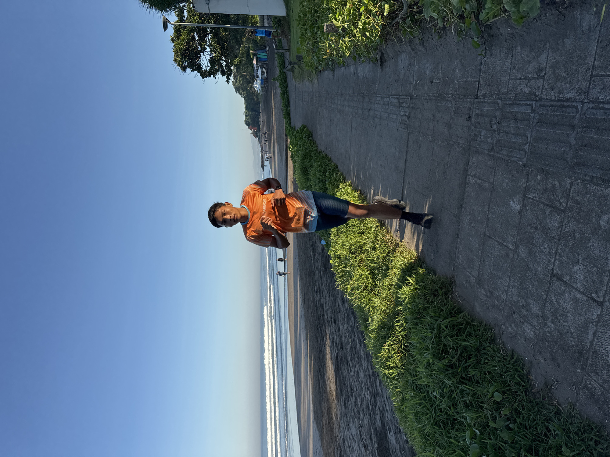 A young man is jogging on a paved pathway adjacent to Kuta Beach in Kuta, Indonesia. He is wearing an orange running shirt and blue shorts. The beach is to his left, with visible waves and a few people in the distance. Lush green vegetation borders the pathway on his right. The sky is clear and blue.
