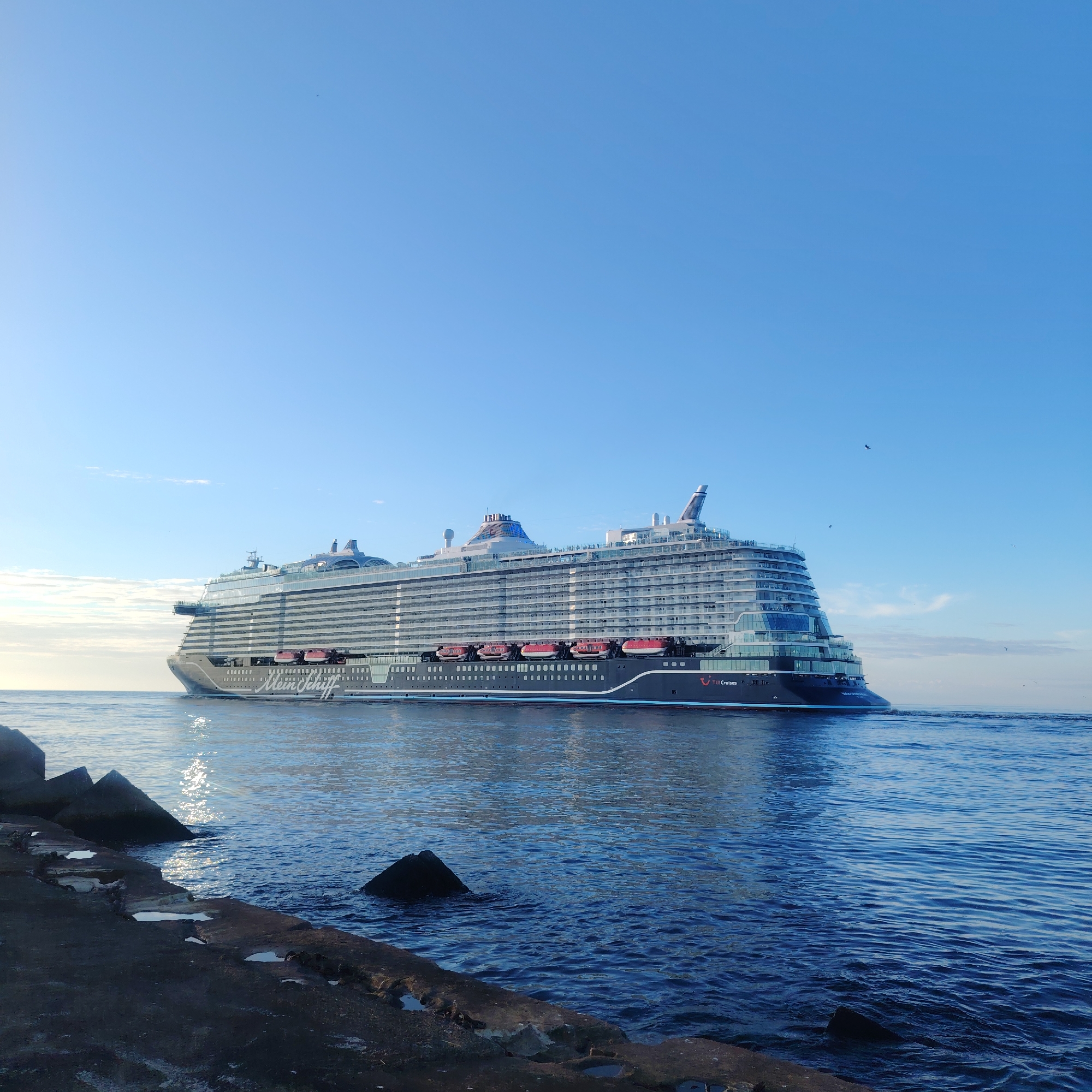 The image shows a large cruise ship, labeled "Mein Schiff", sailing in Riga, Latvia. The ship is predominantly dark in color with red lifeboats along its side. It's moving through a body of water, likely the Daugava River as it approaches the Baltic Sea, as evidenced by the stone breakwater in the foreground. The sky is clear and blue. There are no visible people. The focus is on the ship and the surrounding water and sky.