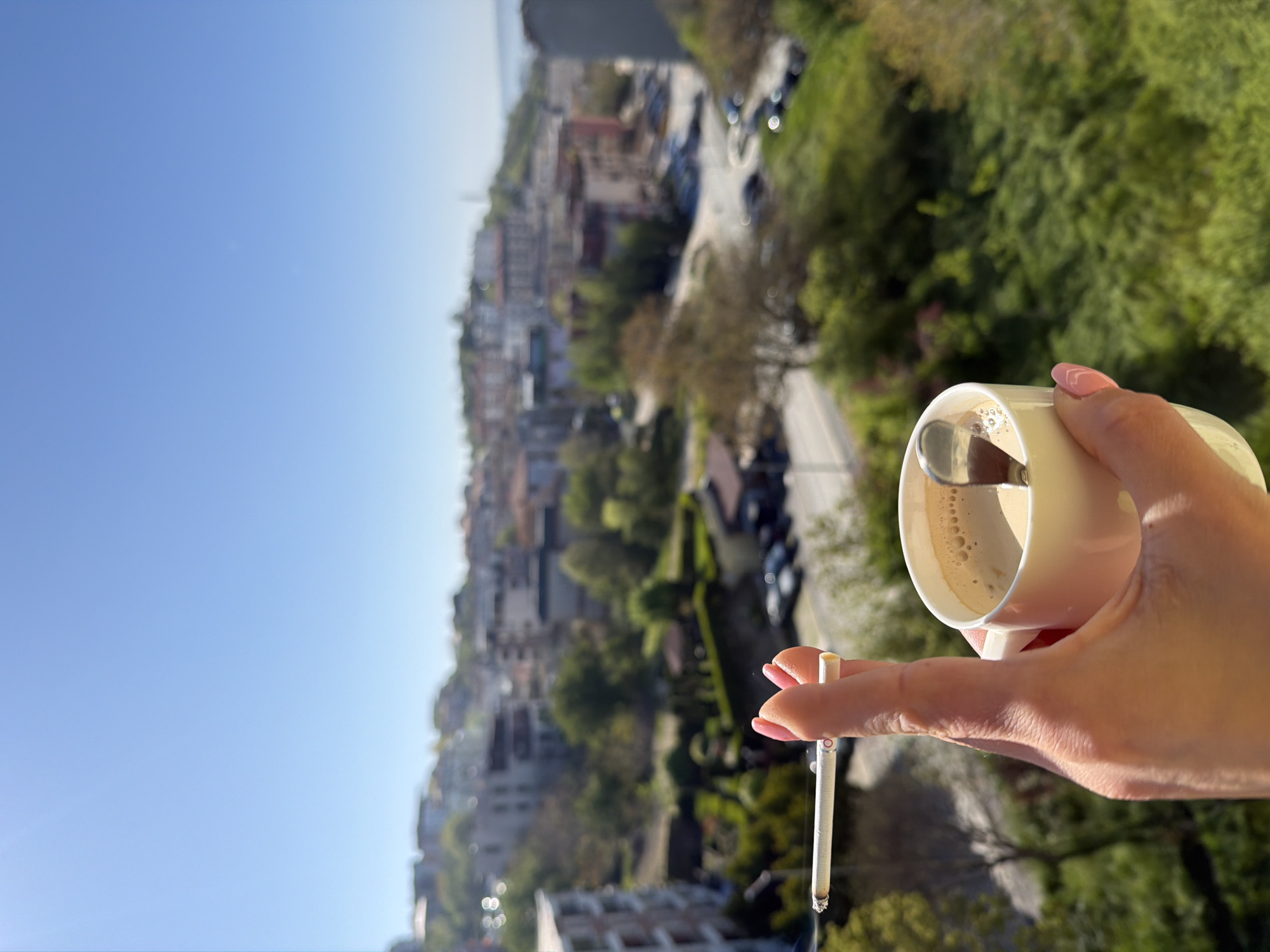 A person is holding a cup of coffee and a lit cigarette on a balcony overlooking a city with green trees, likely in Varna, Bulgaria. The sky is bright blue, suggesting a sunny day. The image is taken from a high vantage point, potentially from a building with multiple floors.