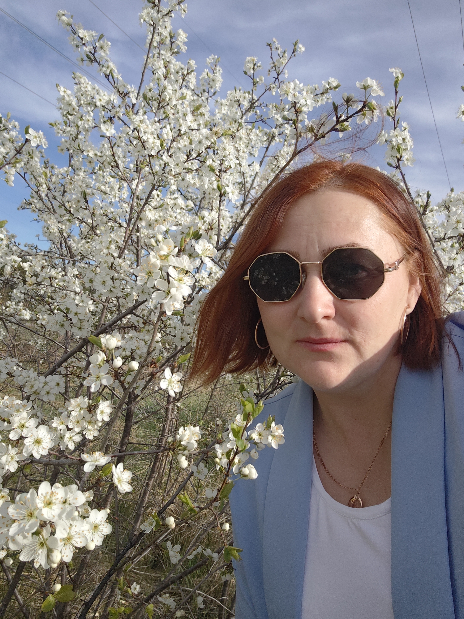 A woman with red hair is standing in front of a flowering tree in Chaikovsky, Russia. She is wearing sunglasses, a blue blazer, and a gold necklace. The tree is covered in white blossoms. The sky is blue with some clouds. There are power lines visible in the background.