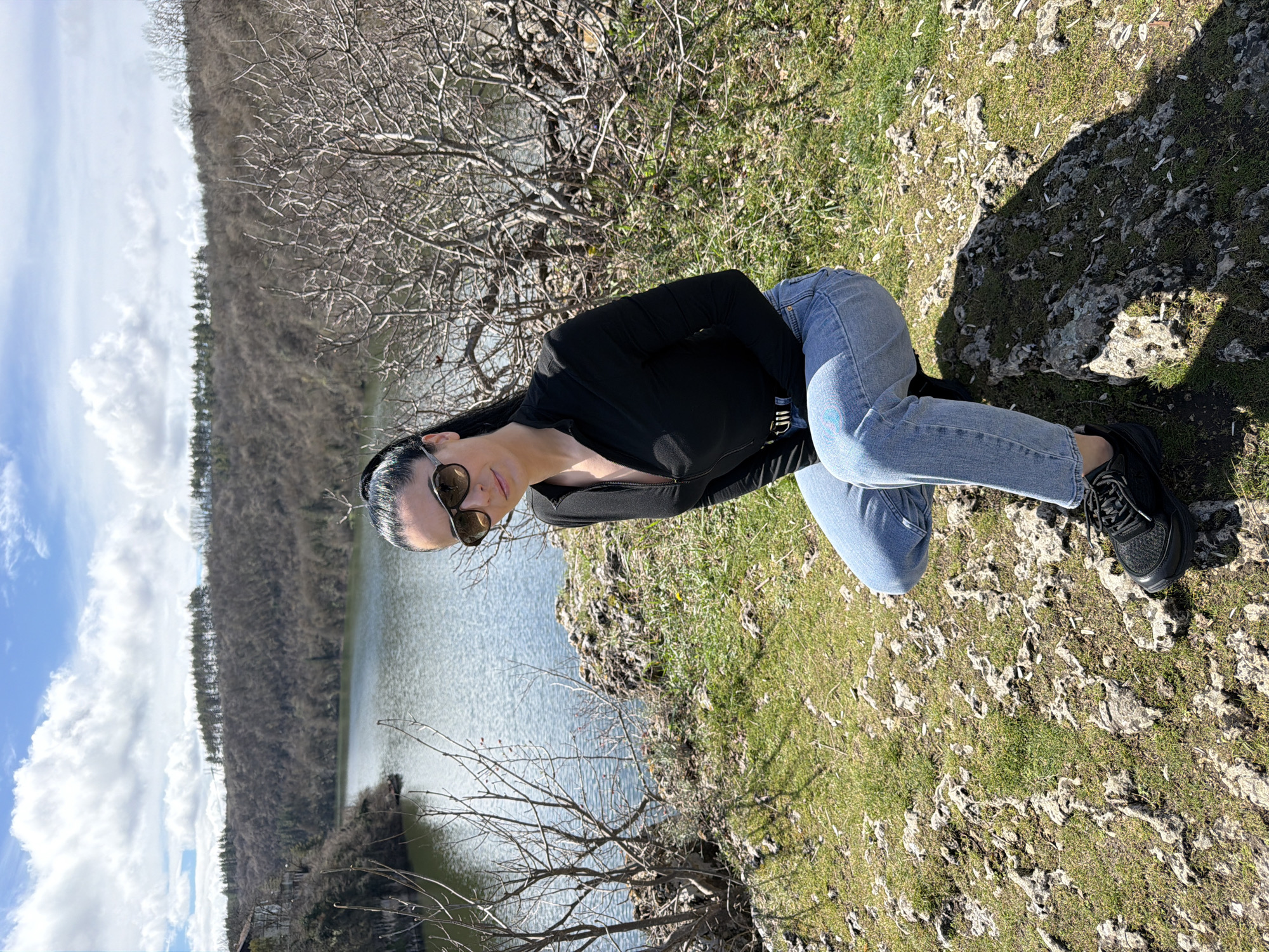 A female subject is observed crouching on a rocky, grassy embankment overlooking a body of water in Kaylaka Park, Pleven, Bulgaria. The subject is positioned slightly left of center, facing the camera with a neutral expression. She wears a black long-sleeved quarter-zip top, light blue denim jeans with a faint light-blue mark on the left thigh, a dark belt with a metallic buckle, and black athletic-style sneakers. Dark sunglasses with prominent frames obscure her eyes. Her dark hair is pulled back into a ponytail.
The immediate foreground consists of rugged, light-colored rocks interspersed with patches of green grass. Bare, leafless shrubbery is present to the subject's left and right. The midground features a calm body of water, likely the Vit River, reflecting the bright, partly cloudy sky. The background shows densely wooded hills with predominantly bare deciduous trees, and a distinct line of evergreen trees visible along the distant ridge. A light-colored building with a dark roof is partially visible on the far left, situated on the hillside near the water's edge. The sky above is blue with scattered white cumulus clouds. The scene is illuminated by natural daylight, casting shadows to the subject's lower right.