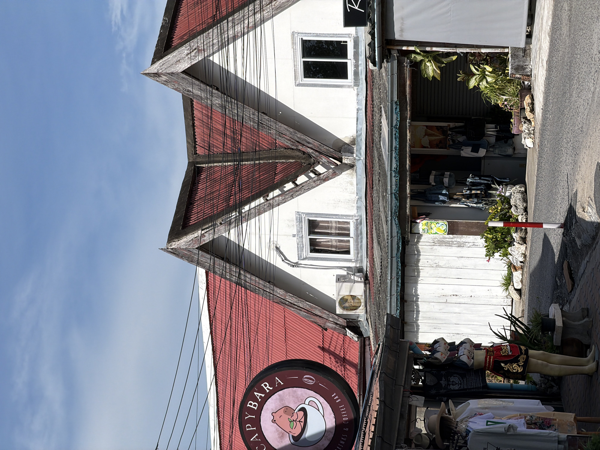 The image depicts a street scene in Baan Plai Laem, Thailand, under a bright, partially clouded sky. The foreground features a paved road and a sidewalk area. On the left, a clothing store displays garments on racks and a mannequin wearing a Muay Thai-style outfit. The mannequin has tan skin and appears to be a headless, partial torso. Behind the store, a building with a distinctive red, corrugated metal roof features triangular gables with weathered wooden beams. The building has white walls and two windows on the upper floor. A round sign for "CAPYBARA DRINKS & COFFEE BAR," featuring a cartoon capybara in a coffee cup, hangs on the left side of the building. An air conditioning unit is visible below one of the windows. To the right, another section of the building has a closed metal shutter and a white awning. Various plants are situated in pots and on the ground around the buildings, and several large rocks are scattered along the sidewalk. Electrical wires are strung across the upper portion of the scene. The overall impression is of a commercial area in a tropical setting, with a mix of retail displays and architectural features characteristic of the region.
