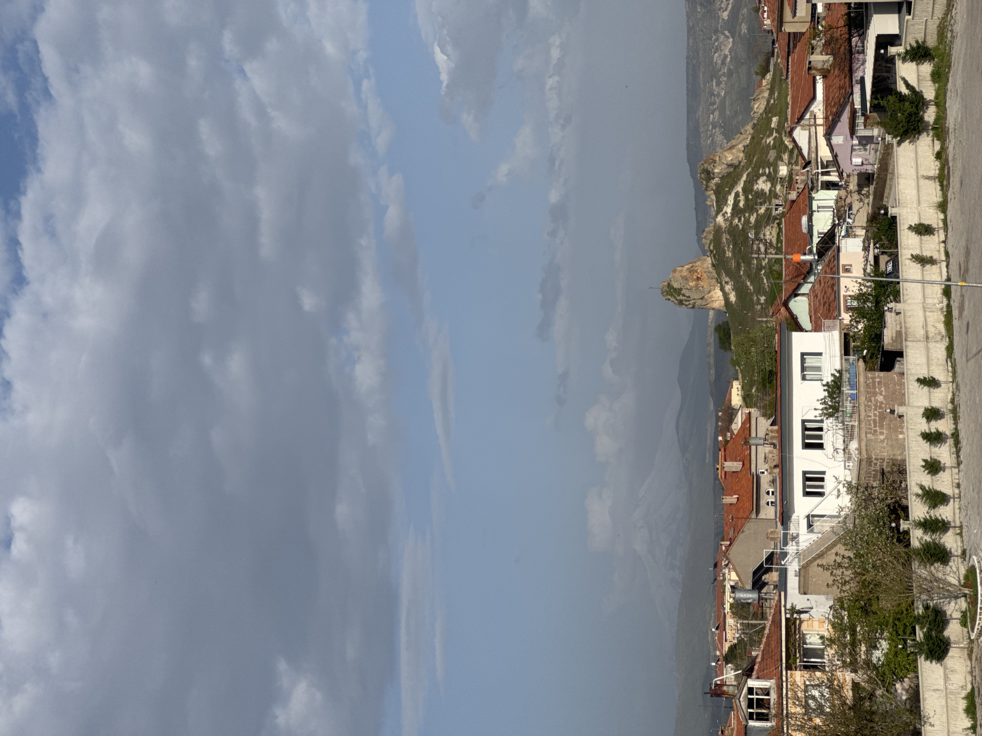 The image presents a view of a hillside town under a partly cloudy sky. In the foreground are the roofs of closely spaced buildings, predominantly red-tiled. A notable rocky outcrop rises behind the buildings, connecting the town to the mountainous landscape in the distance. The architecture features a mix of traditional and modern elements. Adjacent to the buildings is a paved road with regularly spaced, small trees. The town is located in Ürgüp, Turkey, known for its unique geological formations and historical sites.