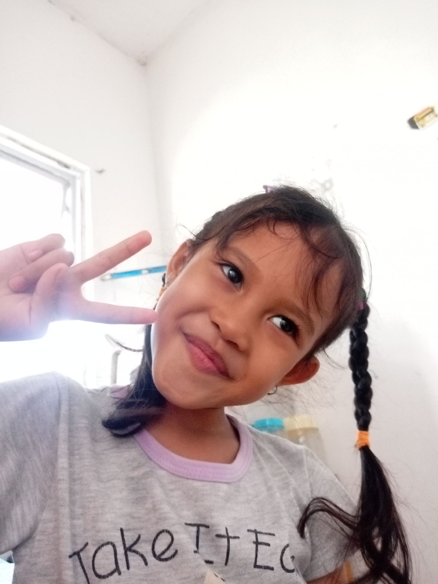 A young girl with dark hair in two braids is shown in a close-up selfie. She is wearing a light gray t-shirt with a lavender collar. She is making a peace sign with her right hand, her fingers held up near her face. The background is a plain white wall with a window on the left side of the image. There are some minor blemishes on the wall. The overall impression is of a casual indoor setting. The image was likely taken in Batam, Indonesia, a city known for its residential areas with simple interior designs.