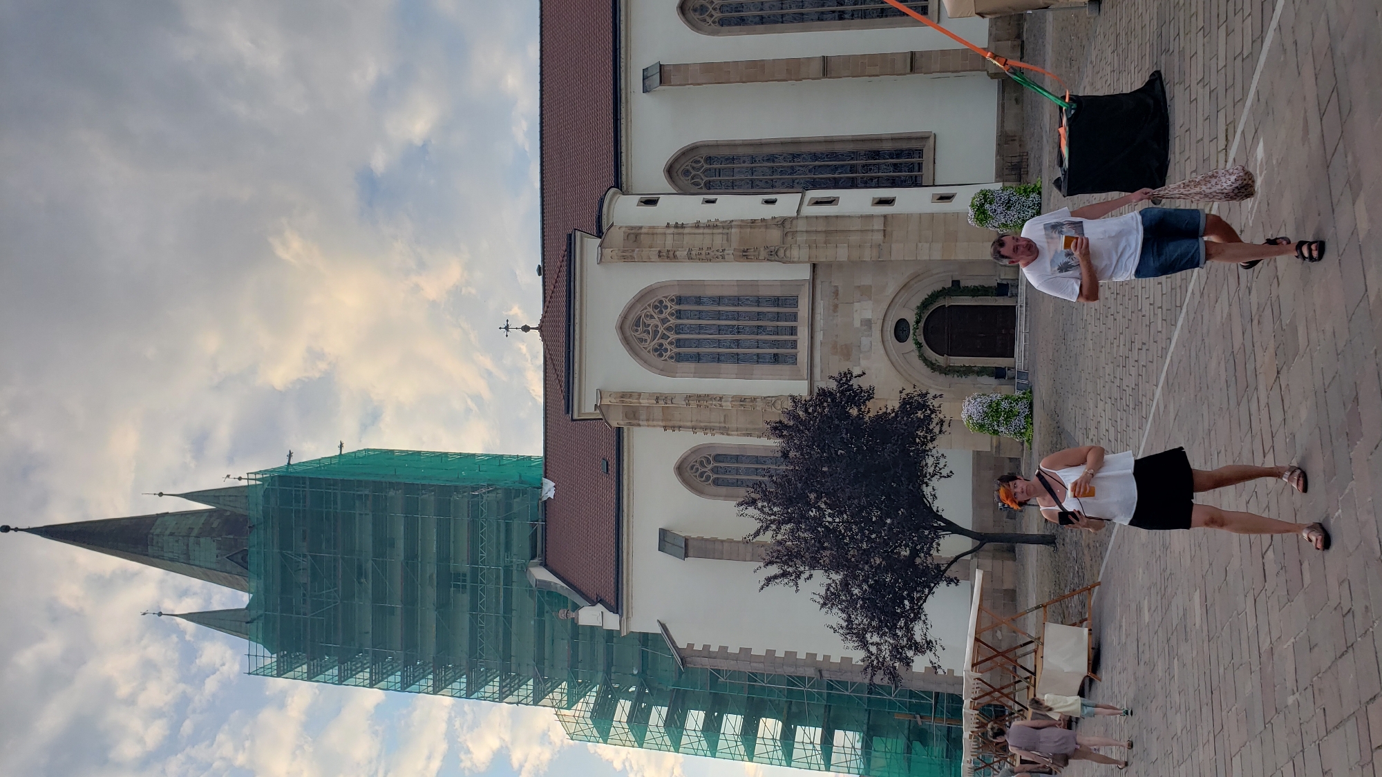 The image shows the interior courtyard of St. Nicholas Cathedral in Prešov, Slovakia. A large, white church building with gothic windows and a red tiled roof dominates the right side of the frame. To the left of the church, a tall bell tower is visible, covered in green scaffolding. In the foreground, a paved courtyard with brickwork is present. Several people are walking through the courtyard, some holding drinks. A woman in a white top and black skirt is in the center, holding a drink. A man in a white t-shirt and denim shorts is to her right, also holding a drink and a shopping bag. In the background, market stalls are set up, and there are decorative floral arrangements near the entrance of the church. The sky is partly cloudy.