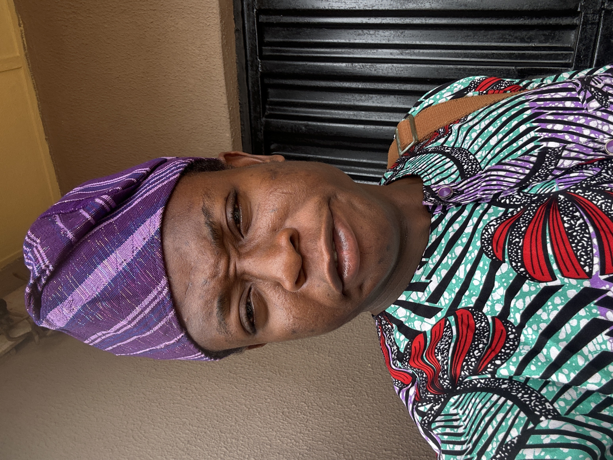 A person with dark skin is visible in an indoor setting. The individual is wearing a purple head covering with striped patterns and a patterned shirt with red, purple, green, black, and white designs, secured with a brown strap across the shoulder. The background features a beige wall on the left and a dark, horizontally-slatted structure on the right. The location is Unknown, Unknown.