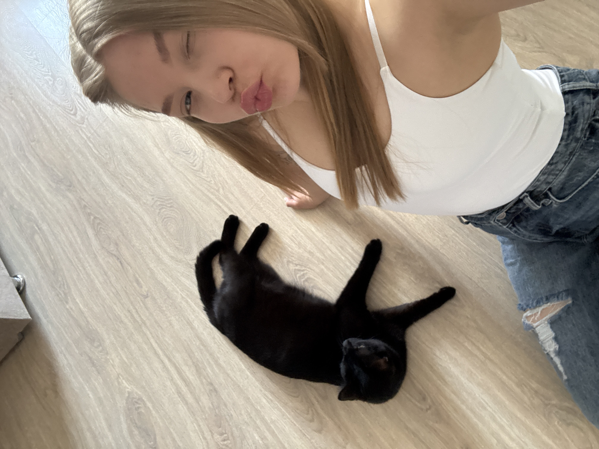 A blonde woman wearing a white tank top and ripped blue jeans is taking a selfie while winking and making a kissing face. She is indoors on a light-colored wooden floor. A black cat is lying on its back nearby. The location is identified as Chaikovsky, Russia.