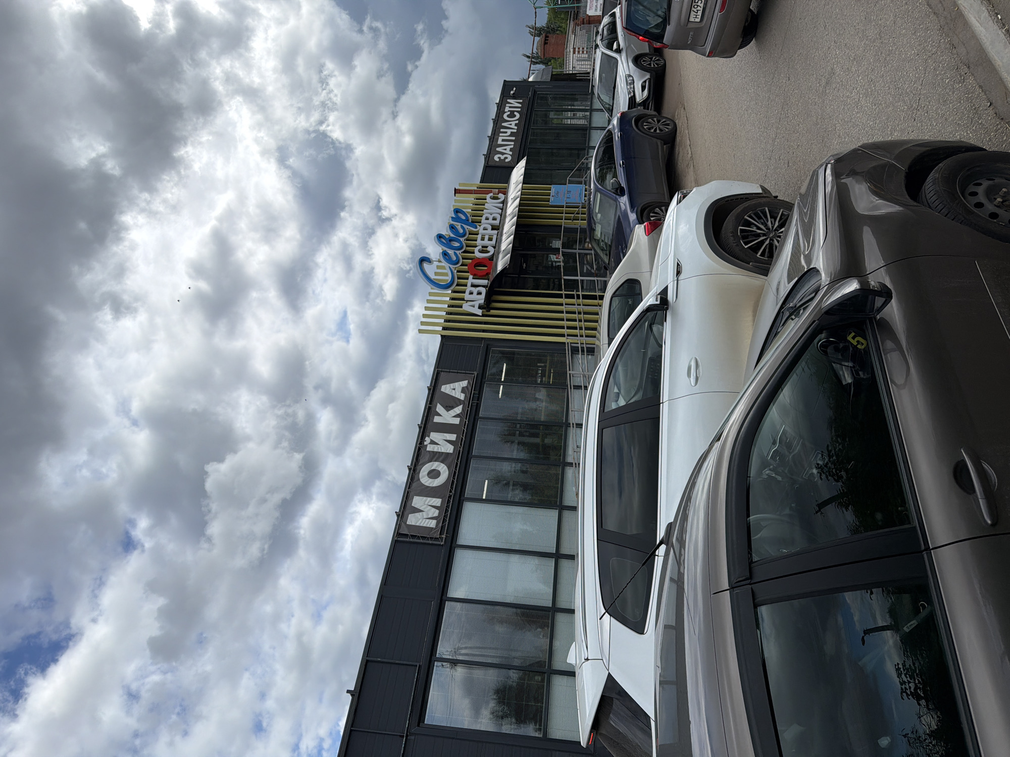 The image captures a street scene in Yelabuga, Russia, featuring a commercial building with prominent signage for "МОЙКА" (Car Wash) and "Север АВТО СЕРВИС" (Sever Auto Service) and "ЗАПЧАСТИ" (Spare Parts). Several cars are parked in front of the building, including a white SUV, a dark blue sedan, a silver sedan, and a gray hatchback in the foreground. The sky above is cloudy. The scene appears to be a typical day in a commercial area.