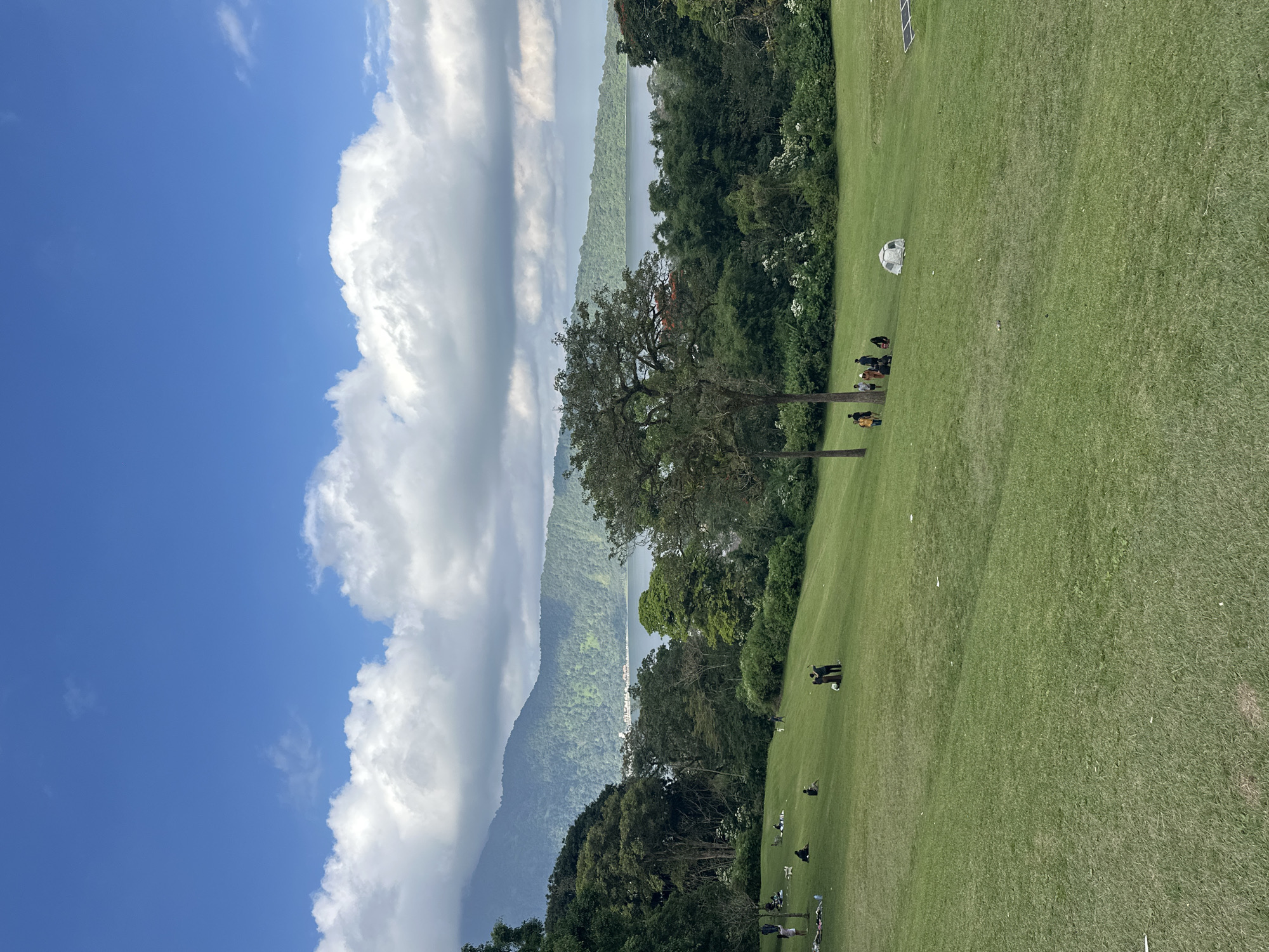 This is a wide-angle outdoor photograph taken on a cloudy day. The scene depicts a large, grassy hillside sloping upwards towards a lake and mountains in the distance. Several people are scattered across the lawn, some sitting, some standing, and a few are gathered in small groups. One group is clustered around a couple of trees in the mid-ground. A small, light-colored tent is visible on the grass further up the slope, and to the right, a solar panel array is partially visible. The sky is bright blue with large, puffy white clouds. The vegetation is lush and green, with trees framing the lake and the lower slopes of the mountains. The overall impression is of a peaceful, recreational outdoor setting, likely a park or scenic overlook. This location is identified as being in Baturiti, Indonesia.