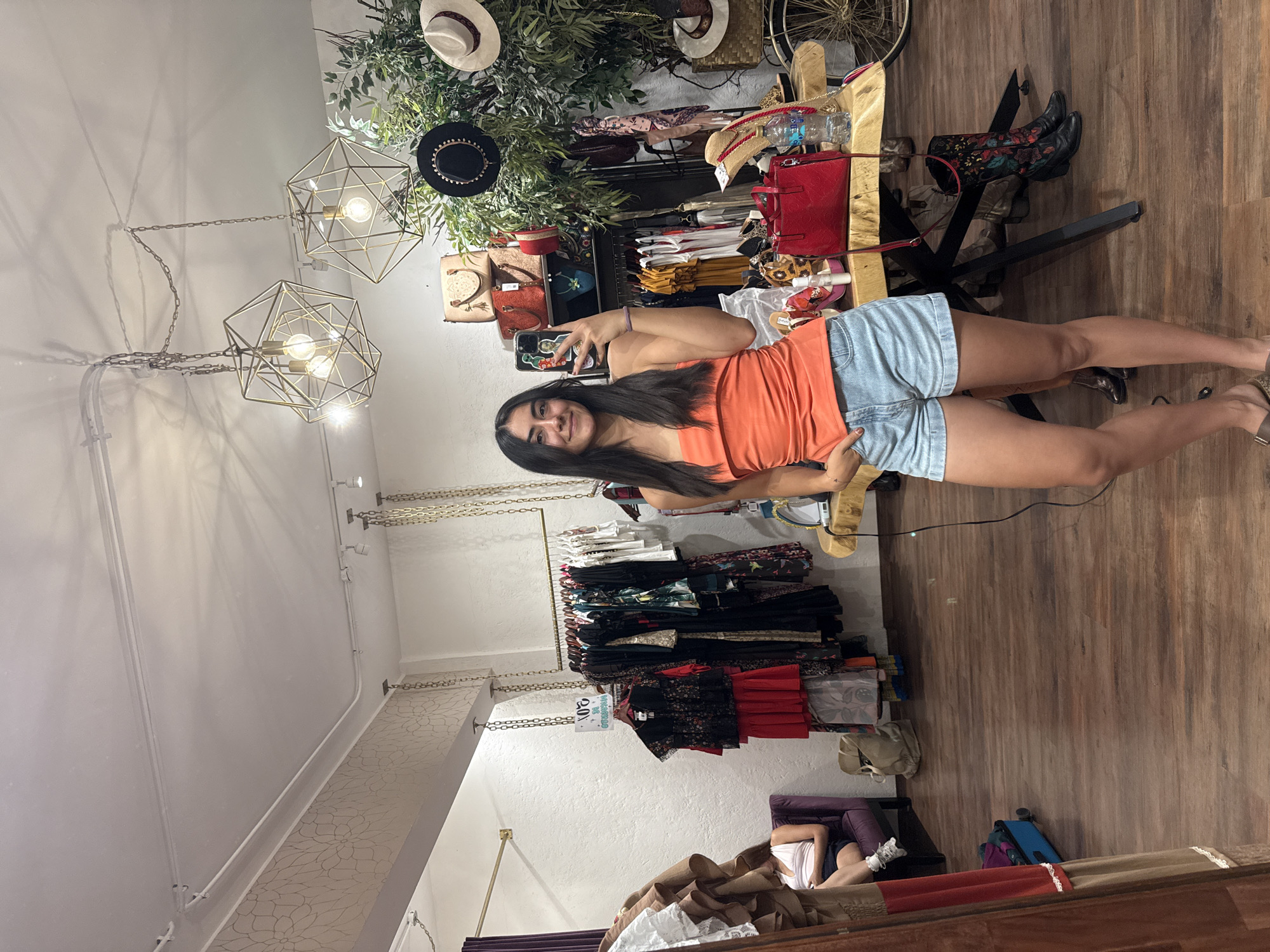 A young woman stands inside a retail shop in Domingo Arenas, Mexico, taking a selfie. She is wearing an orange top and light blue denim shorts. The store's interior features clothing racks suspended by chains, displaying a variety of dresses and tops. Above the clothing racks are shelves decorated with hats, plants, and handbags. Geometric light fixtures hang from the ceiling. The floor is made of wood. Other objects include a table with merchandise on top, a chair, a suitcase, and a reclined person on a chair to the bottom left corner of the image.