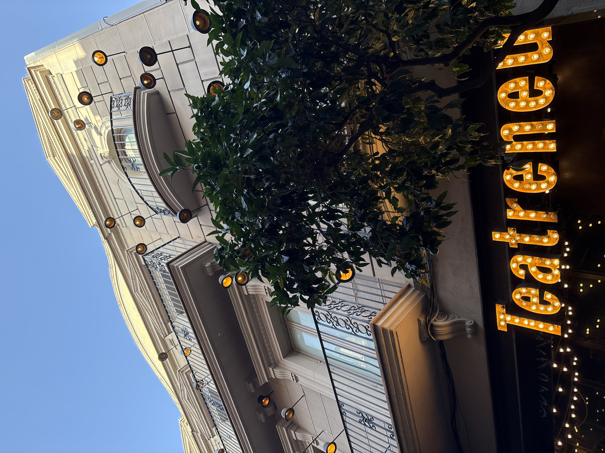 The image shows a street-level view of a building in Barcelona, Spain, at dusk or early evening. The building facade is light-colored and features balconies with decorative metal railings. Several outdoor lights, resembling vintage bulbs on articulated arms, are mounted on the facade. A large, leafy tree partially obscures the building's lower floors and the sign below. The sign, illuminated with warm, glowing bulbs, spells out "Teatrenou." String lights are also visible below the main sign. The sky above is a clear, deep blue. The scene conveys a lively, theatrical atmosphere, suggesting the entrance to a performance venue or a place of entertainment.
