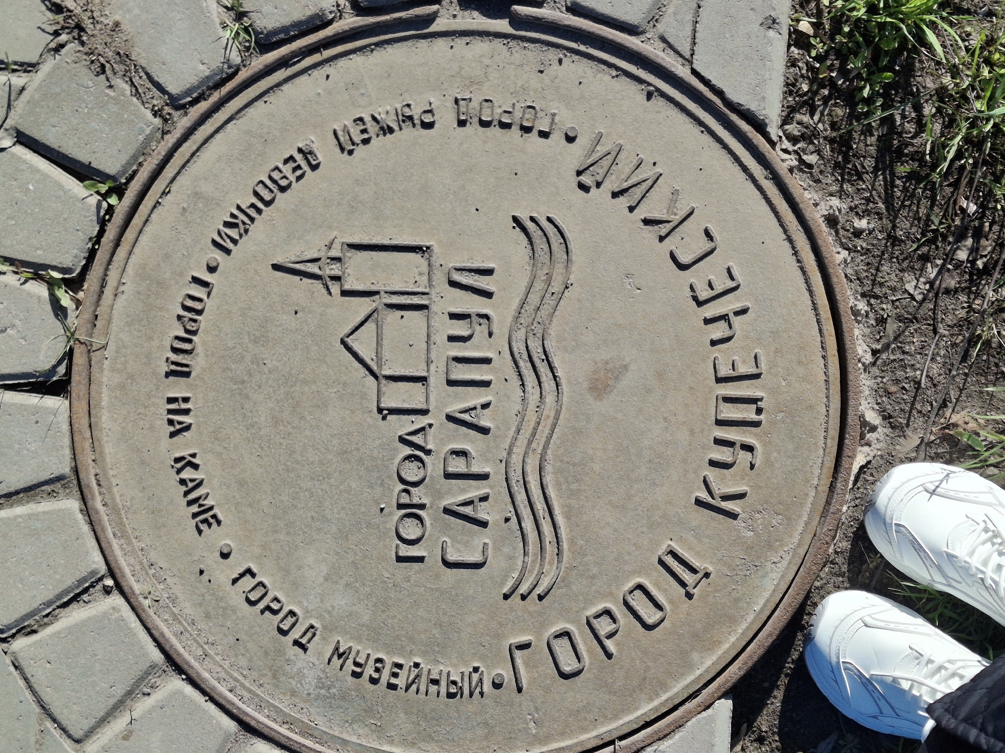 An overhead shot captures a circular, weathered metal manhole cover set into a paved surface, partially surrounded by dirt and grass. The manhole cover features raised Russian text and a central emblem, indicating its location in Sarapul, Russia. The emblem depicts a stylized building, resembling a tower or lighthouse, beside three wavy lines, likely symbolizing the Kama River.
The text on the cover includes "ГОРОД САРАПУЛ" (CITY SARAPUL) in the center. Arranged in an arc around the perimeter, the text lists various epithets for the city: "КУПЕЧЕСКИЙ • ГОРОД" (MERCHANT • CITY), "МУЗЕЙНЫЙ • ГОРОД" (MUSEUM • CITY), "ГОРОД НА КАМЕ" (CITY ON KAMA), and "ГОРОД РЫЖЕЙ ДЕВОЧКИ" (CITY OF THE RED-HAIRED GIRL).
The manhole cover has a rusty rim and is surrounded by grey concrete pavers on the left and bottom. To the right and top, the ground transitions to dry soil with patches of green grass and weeds. Strong daylight indicates a clear, sunny day, with subtle shadows visible from the grass and across the cover. In the bottom right corner, a person's feet wearing clean white sneakers are visible, suggesting someone standing near or over the manhole.