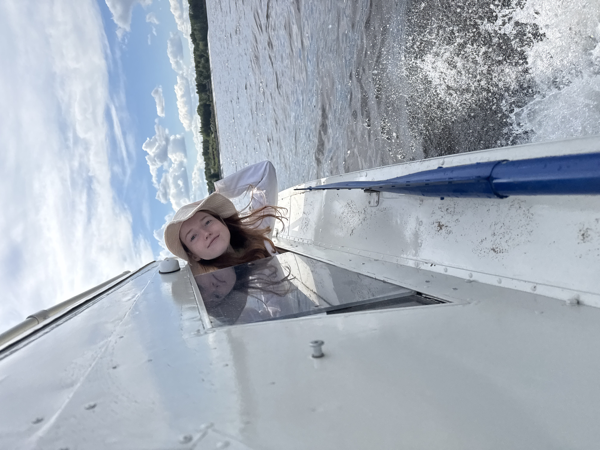 A daytime image, rotated counter-clockwise by 90 degrees, depicts a young person on a moving vessel on a large body of water near Chaikovsky, Russia.
The central subject is a young girl or woman with long reddish-brown hair and fair skin, wearing a light-colored hat and shirt. She is seen from the chest up, looking out with a gentle smile from what appears to be a window or hatch of a white boat or hovercraft. Her expression suggests enjoyment of the ride.
The vessel is actively moving, generating a significant spray of white water next to its side, indicating considerable speed. A blue rail or bar is visible along the side of the white vessel structure. In the background, a wide expanse of water stretches towards a distant forested shoreline, dense with green trees. The sky above is bright blue with scattered white, cumulus clouds. The lighting suggests a clear, sunny day, likely in the late morning or afternoon. No visible text is present in the image.