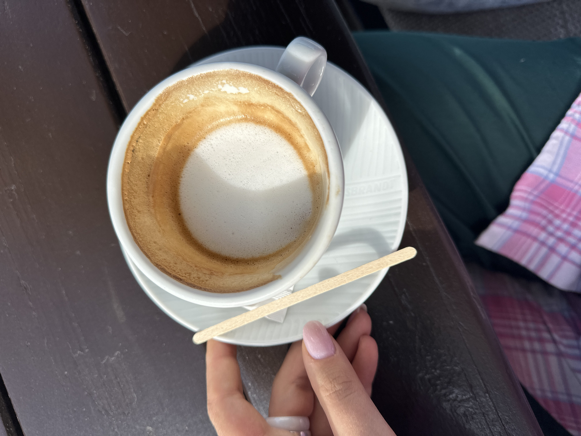 An overhead shot shows a partially consumed cappuccino in a white cup and saucer on a brown wooden table. A light-colored wooden stirring stick lies across the saucer. A person's hand, with a light pink manicure and a ring, touches the saucer. In the background, there is a green cushion and a pink and white checkered fabric. The location is Šušanj, Montenegro.