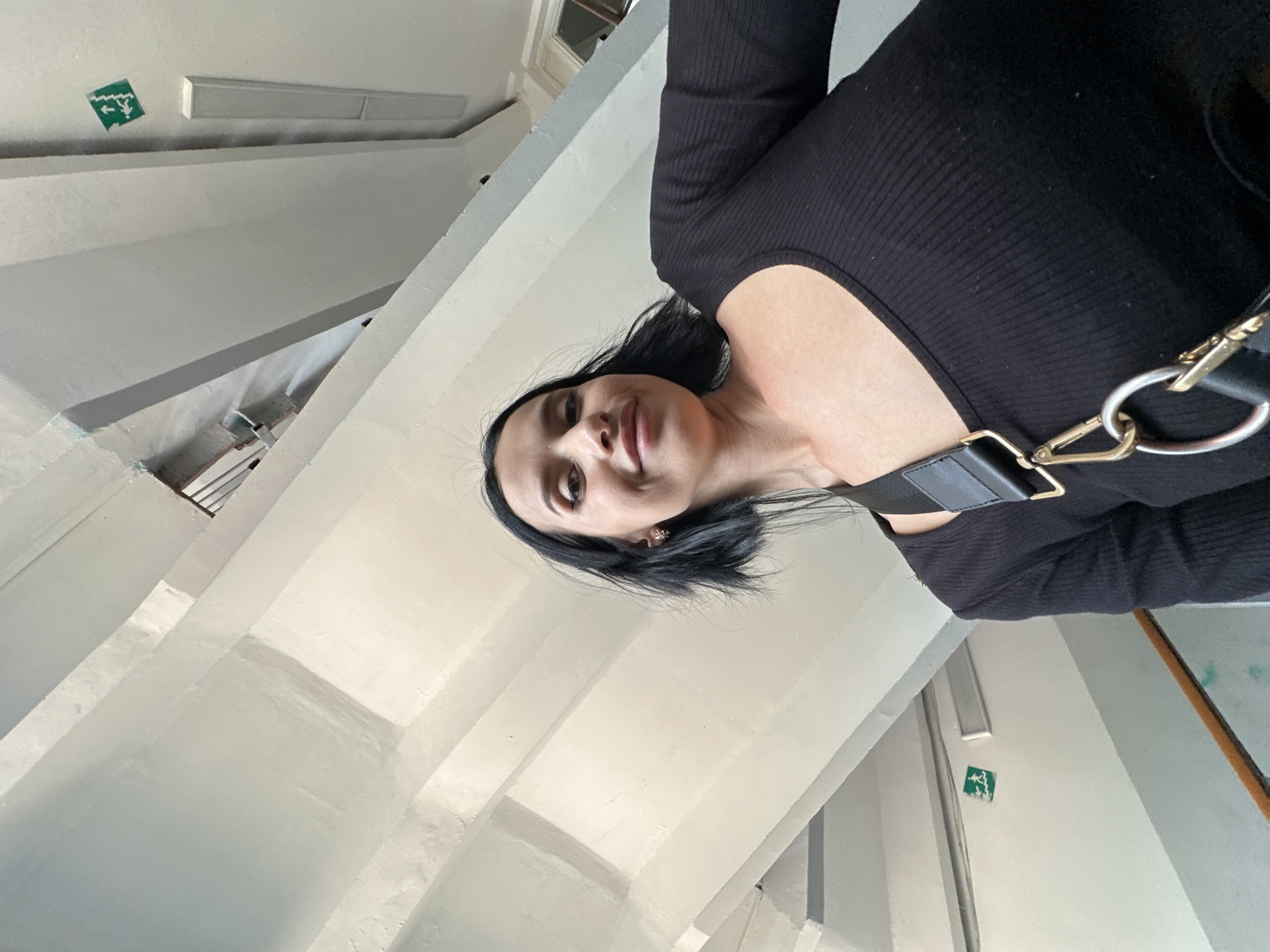 A young woman with dark hair and a black top is taking a selfie on a staircase. The staircase appears to be inside a building with white walls and ceilings, and metal railings. There is a green exit sign on the wall. The lighting suggests it is daytime.