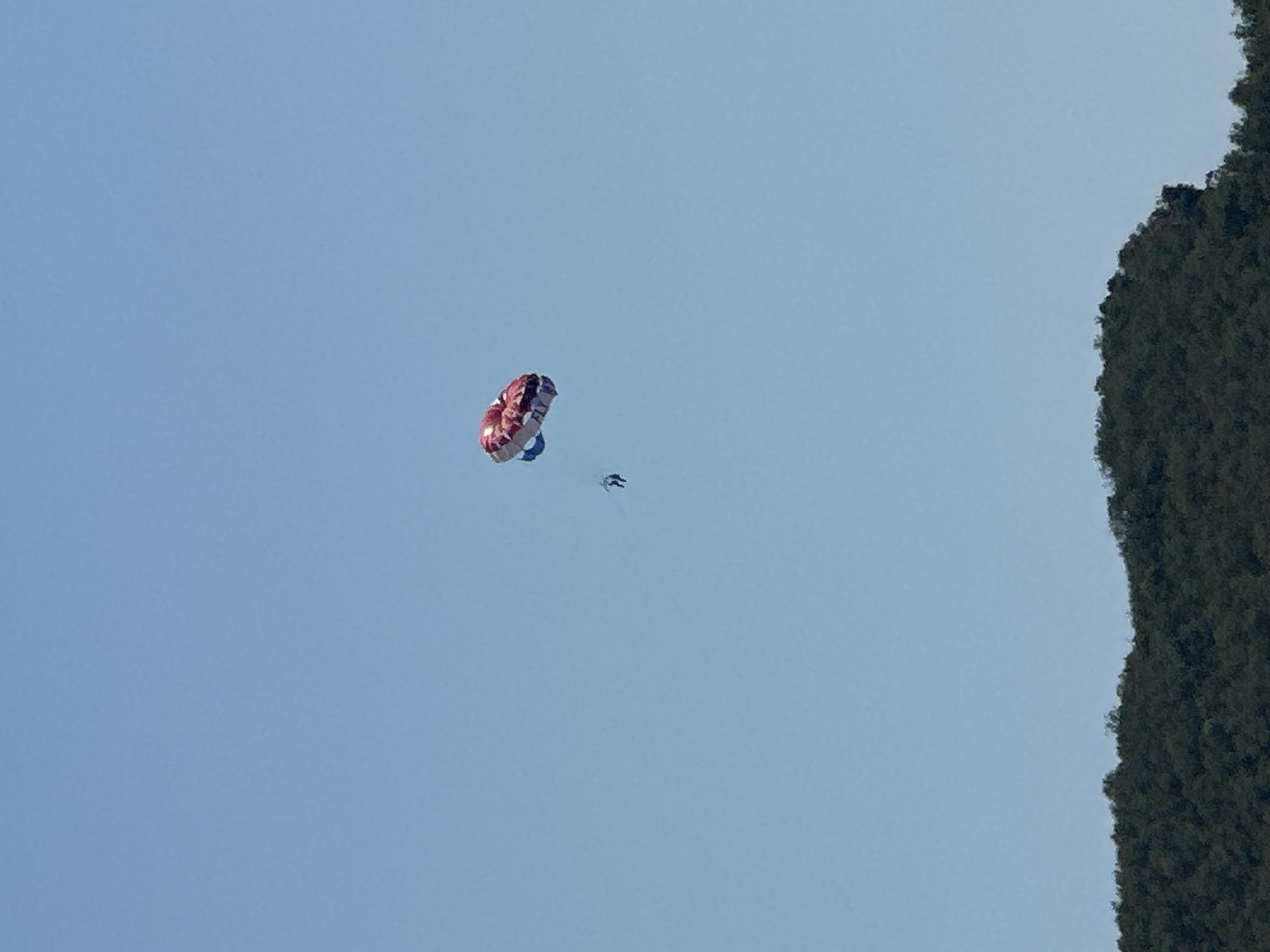In the clear blue sky above Budva, Montenegro, two figures engage in parasailing. A brightly colored parasail, predominantly red and white with blue accents, floats in the upper right quadrant. Below it, a single person, dressed in dark clothing, is suspended from a tow rope. Further down and to the left, another person is also visible, trailing behind. In the lower portion of the image, the dark green, densely forested slopes of a hillside form the horizon. The scene is calm, with no visible boat or watercraft to which the parasailers are attached, implying they are in mid-flight.