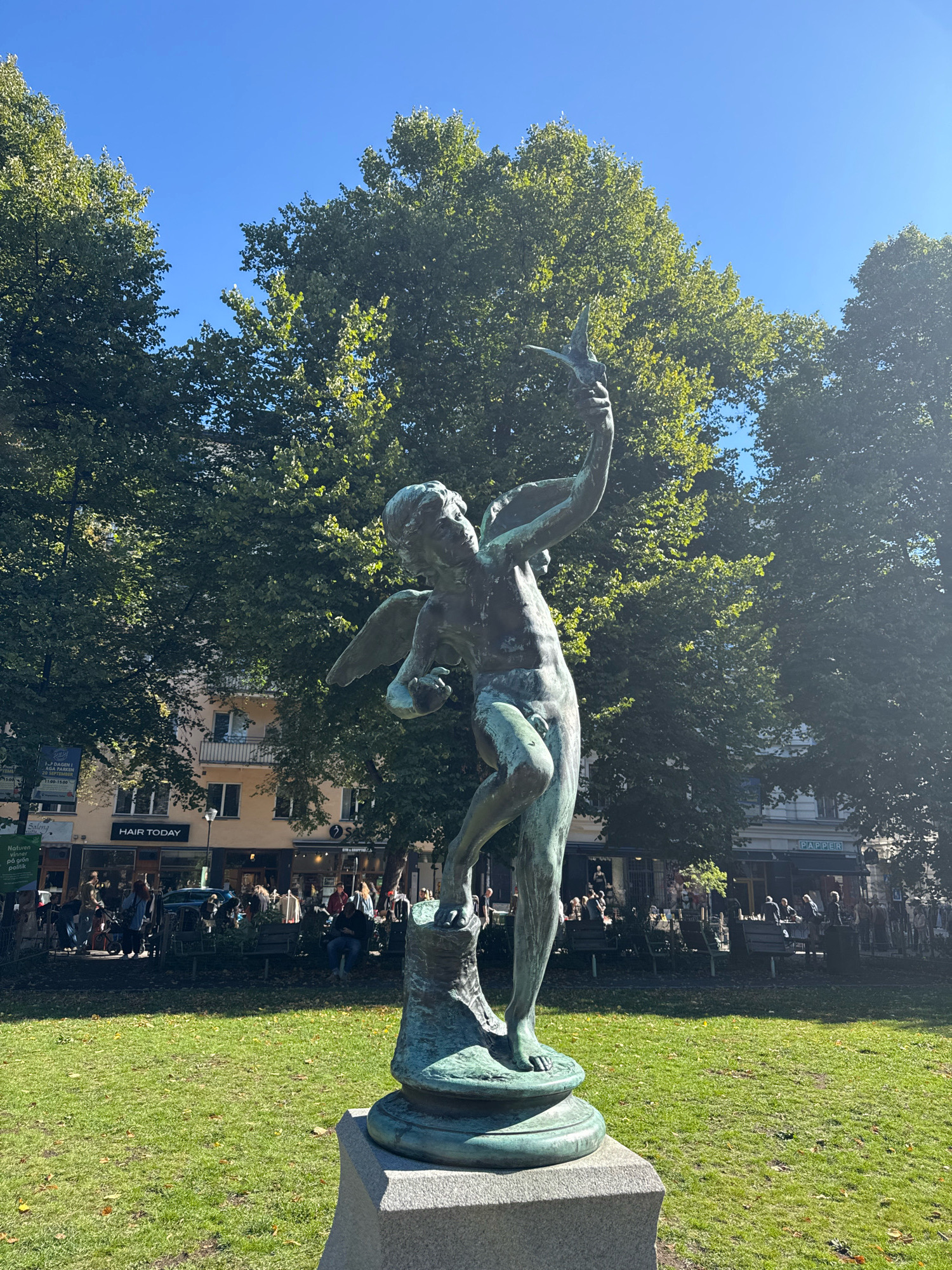 In Stockholm, Sweden, a bronze statue of a winged cherub stands in a park under a clear blue sky. The statue is positioned on a stone pedestal, with the cherub holding a bird in its outstretched hand. The cherub is depicted with its body angled to the right, its left arm raised, and its right arm holding the bird. Green grass covers the ground around the pedestal, and large trees with dense foliage form a backdrop. In the background, there are buildings with windows and balconies, and several people are visible in the park, some sitting on benches and others walking. Shop signs are visible on the buildings, including "HAIR TODAY" and "PAPER". The lighting suggests it is daytime, with sunlight casting shadows on the grass.