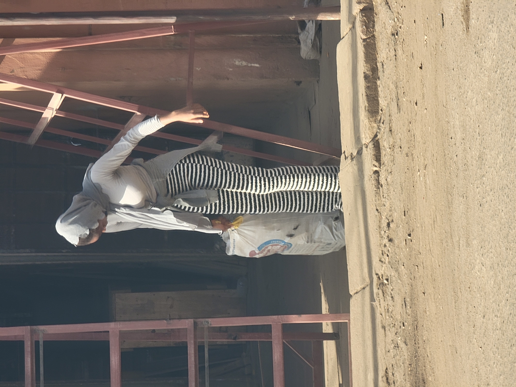 This vertical-oriented image captures a daylight scene at what appears to be a construction site in Maiburiji, Nigeria. The central subject is a person, likely female, wearing a light-colored long-sleeved top, a matching head covering, and striking black and white striped trousers.
The individual is positioned within a structure defined by red-painted metal scaffolding or framework, which is visible in both the foreground and background. The person is bent over, with arms extended, seemingly interacting with a large white bag on the ground. This bag features blue and red graphics, and the word "Plaster" is clearly visible on its side, indicating it contains a building material. A small yellow object is also partially visible near the top of the bag.
To the right of the frame, a roughly textured, light-colored concrete wall stands prominently. The scene is illuminated by natural light, confirming it is daytime. The combination of the metal framework, the concrete wall, and the bag of plaster strongly suggests an ongoing construction or renovation project where the individual is engaged in some form of labor.