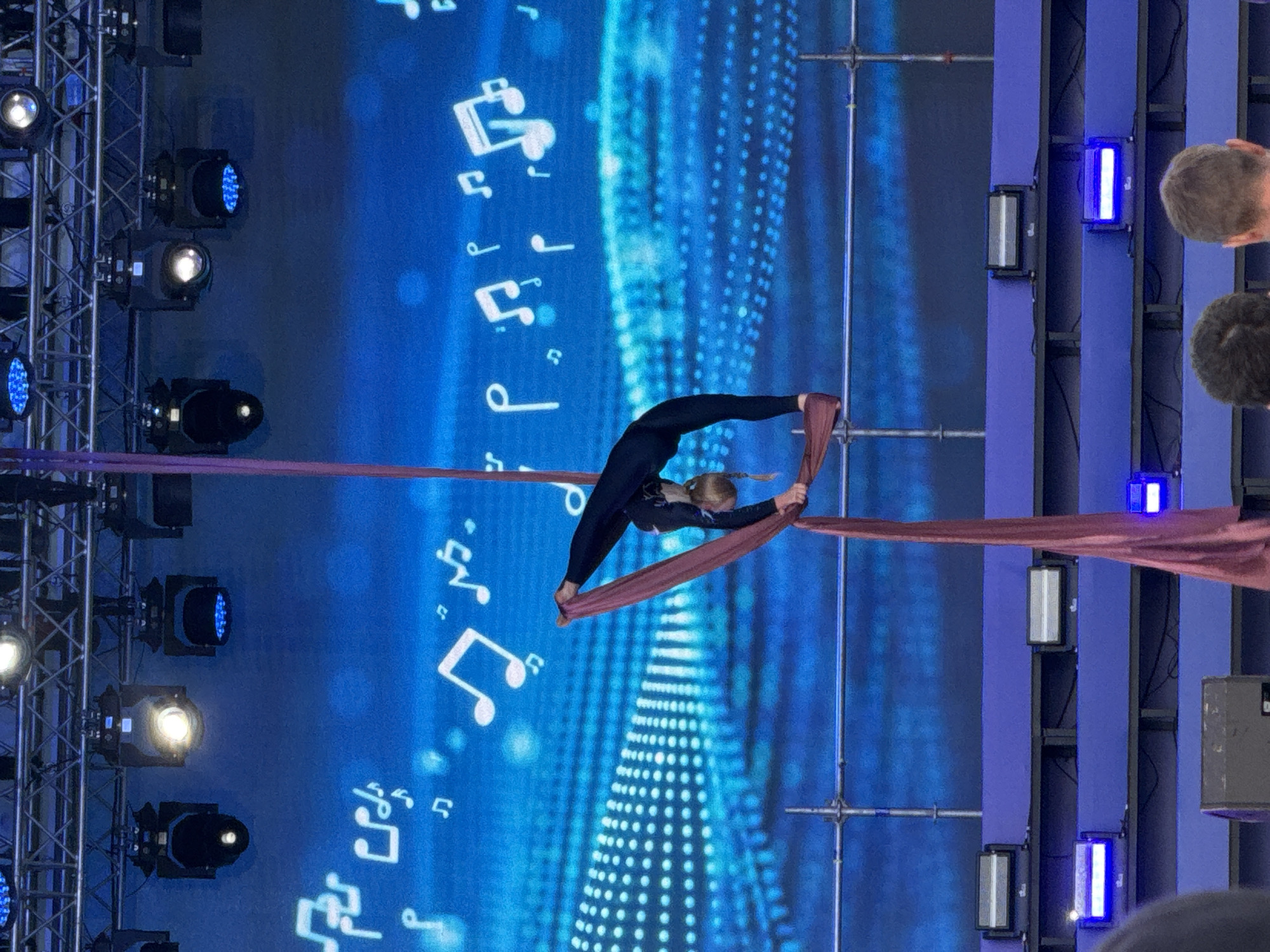 In Izhevsk, Russia, an aerial performer is captured mid-air during a performance. The performer, dressed in a black full-body suit, is suspended by a maroon fabric aerial sling. They are performing an acrobatic pose, with their body arched backward and their legs extended, one foot resting on the sling. The background features a vibrant blue screen displaying a pattern of white musical notes and blurred lights, suggesting a stage or concert setting. Above the performer, a truss structure holds multiple spotlights, some of which are illuminated. Below the stage, rows of blue and white stage lights are visible, illuminating the platform. The image is taken from a slightly low angle, focusing on the aerial artist and the stage setup.