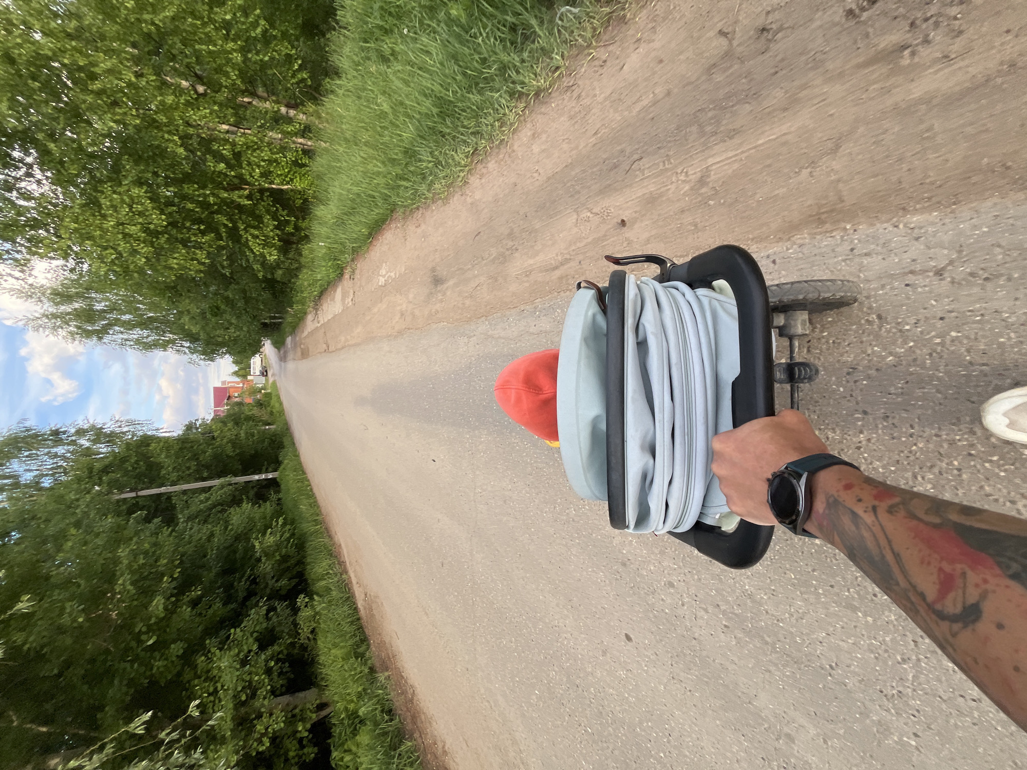 A first-person perspective image depicts a person walking along a dirt road in Chaikovsky, Russia, while pulling a stroller containing a child. The road stretches into the distance, bordered by green vegetation on both sides. The person's arm, adorned with tattoos and a smartwatch, is visible holding the stroller's handle. The stroller is black and contains a folded light blue or grey fabric. A child's red hat is also visible inside the stroller. The background features trees, a partially visible blue sky, and buildings in the distance.