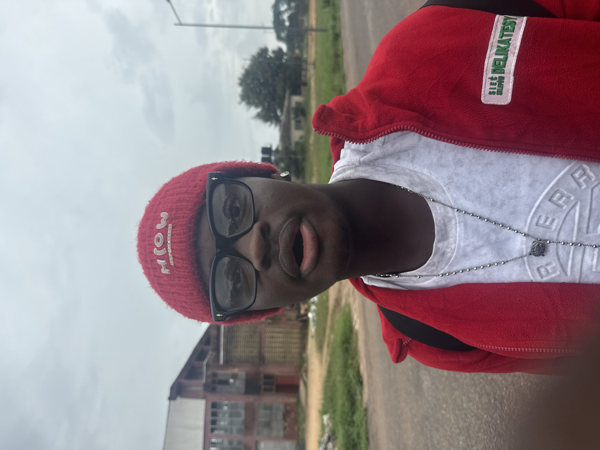 A young Black male wearing a red knitted beanie with "HIOW" embroidered in white, black-rimmed glasses, and a red zip-up jacket over a white t-shirt stands outdoors. He is wearing a silver chain necklace. The background features a weathered, reddish-brown building with visible windows and a dark red door, and a light gray, cloudy sky. To the right, a street lamp and trees are visible, suggesting a semi-urban or rural setting in Ihiagwa, Nigeria. The ground is paved with asphalt, and there is a patch of green grass to the left of the frame.
