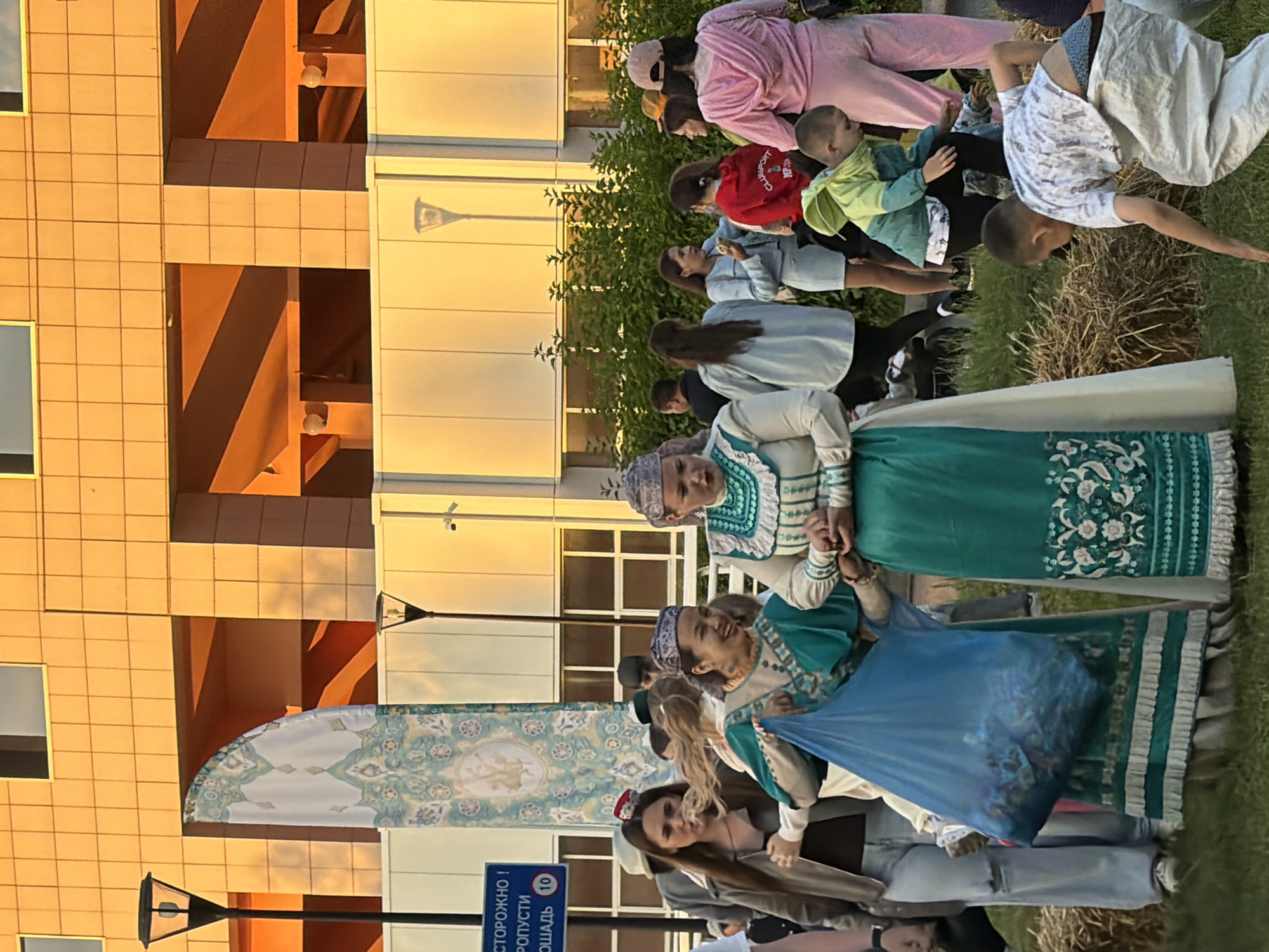 In the city of Unknown, Unknown, a group of people are gathered outdoors, possibly for a cultural event or festival, as indicated by the traditional clothing worn by some individuals. The setting appears to be in front of a modern building with orange-tinted windows and architectural features. A large banner with intricate patterns is visible on the left side of the frame. In the foreground, two women are dressed in elaborate, traditional attire. One woman in a teal and white dress smiles broadly while holding a large blue plastic bag filled with items. The other woman, also in a similar traditional outfit, is looking to her right. A young boy in a green jacket and another in a white shirt are seen in the middle ground, interacting with hay bales. Several other individuals, dressed in casual modern clothing, are observing or participating in the event. The lighting suggests it is either early morning or late afternoon, with a warm glow from the sun illuminating the scene. The ground is covered in grass and hay, further suggesting an outdoor event with rustic elements. A street light is visible on the left, and another in the background.