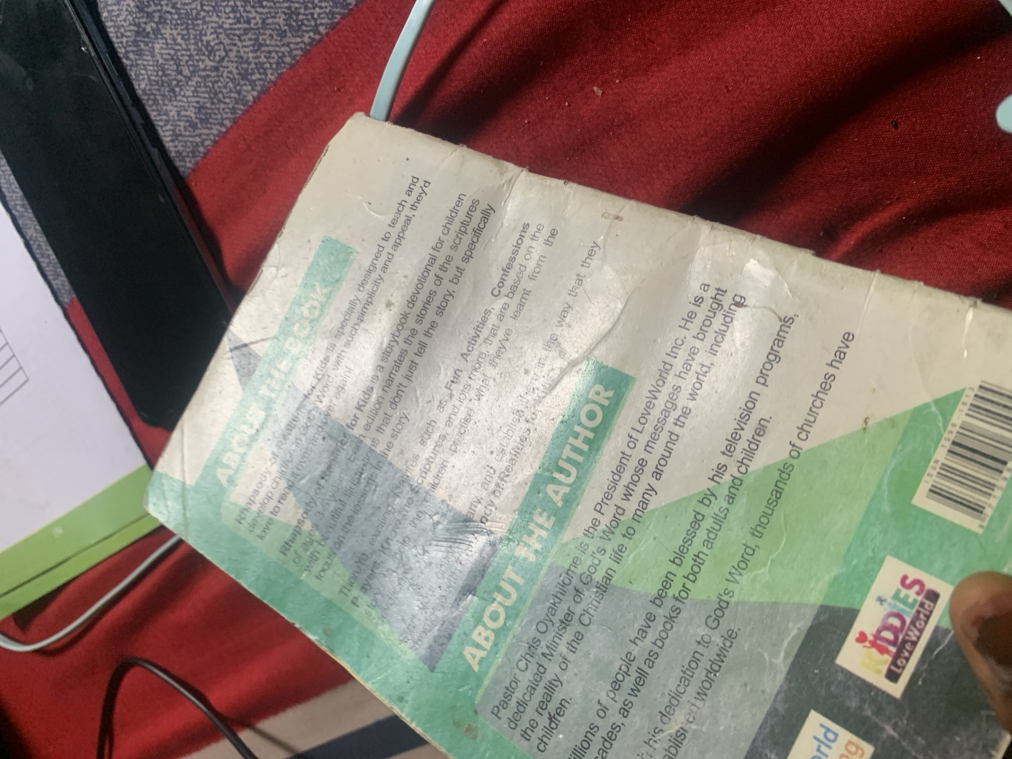 A close-up, high-angle shot shows the back of a tattered children's book. The book's cover, made of a light blue material, is folded over a dark red fabric surface. The text on the book is primarily in English, with titles such as "ABOUT THE BOOK" and "ABOUT THE AUTHOR" visible. The book appears to be a devotional for children, featuring stories, scriptures, and prayers. A logo for "KIDDIES LOVEWORLD" is present on the lower left portion of the book. The book's condition suggests it has been used extensively. The background fabric is a rich, deep red, with visible folds and texture. Several black cables are also visible, partially obscuring the book and the red fabric. The lighting is artificial and casts shadows. There is no information within the image to indicate a specific location in Jauro Yinu, Nigeria.