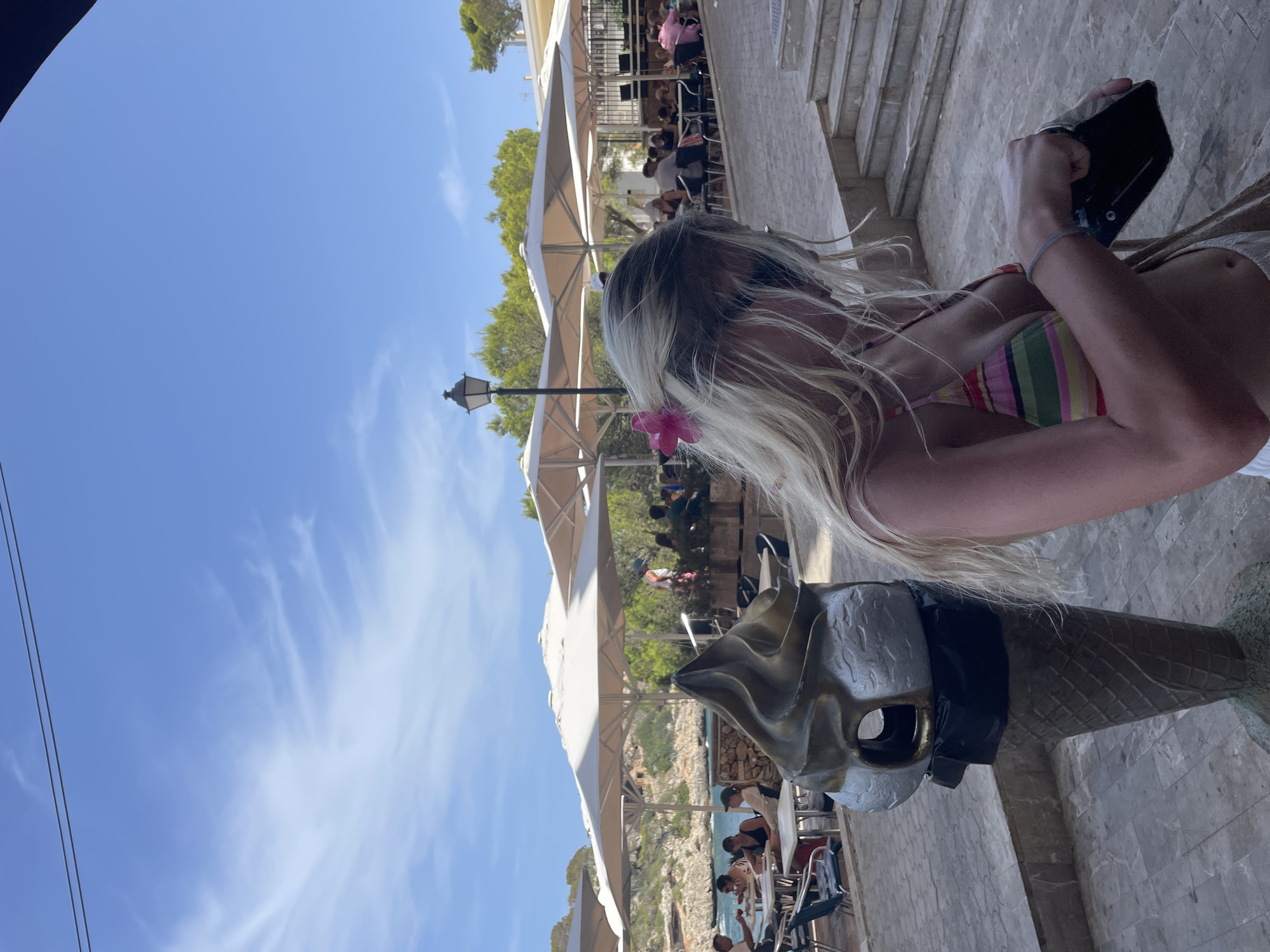 The image shows a woman in a striped bikini and white shorts standing outdoors. She has blonde hair with a pink flower tucked behind her ear. She is holding a black object in her hands. To her left, there is a sculpture of an ice cream cone that appears to be a trash can. In the background, there are tables and chairs set up under large umbrellas, and people are seated at the tables. Beyond the seating area, there is a body of water and a rocky coastline with trees. The sky is blue with some wispy clouds. The scene appears to be a waterfront cafe or restaurant, likely in a Mediterranean coastal town. This location is the municipality of Santanyí, Spain, specifically a coastal area, possibly near Cala d'Or.