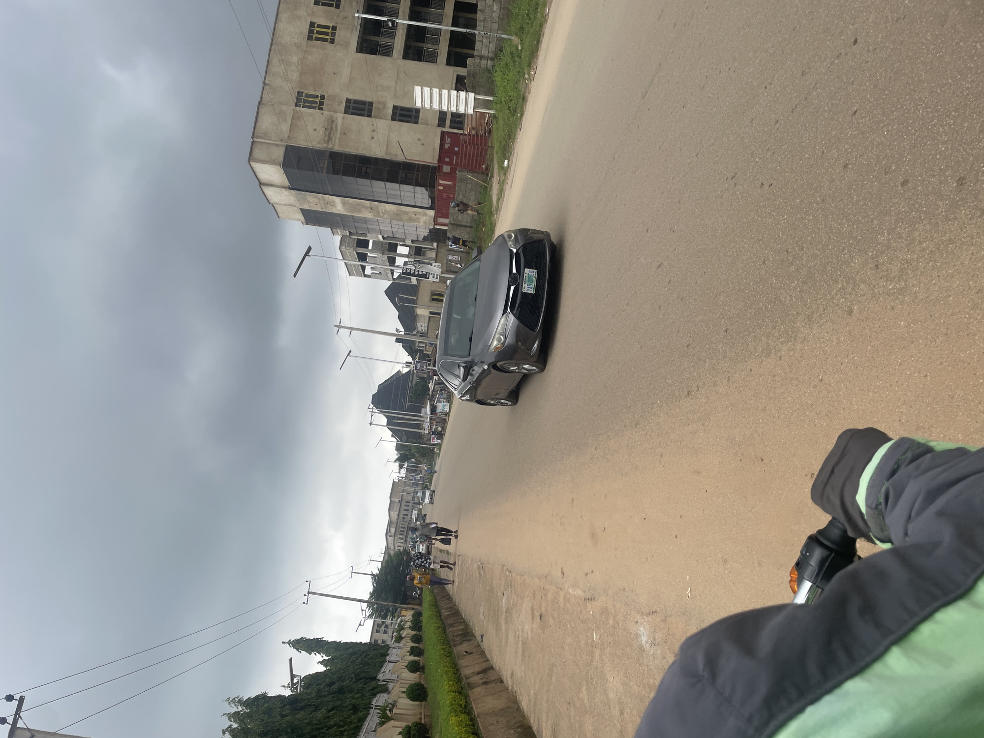 The image captures a street scene in Abuja, Nigeria, under an overcast sky. In the foreground, the perspective is from someone riding on a motorcycle, with a portion of their jacket visible on the left. The road ahead is paved with asphalt and appears to be a two-lane street. On the left side of the road, a paved walkway runs alongside a well-maintained green lawn bordered by a concrete curb. Beyond the lawn, there are buildings and trees.
Several individuals are walking on the walkway. Further down the road, a dark gray sedan is in motion, with its license plate visible. The buildings in the background are a mix of styles, including modern structures with glass facades and what appear to be older, more utilitarian buildings. There are also power lines and streetlights visible along the road. The overall impression is of an urban environment in the midst of activity.
