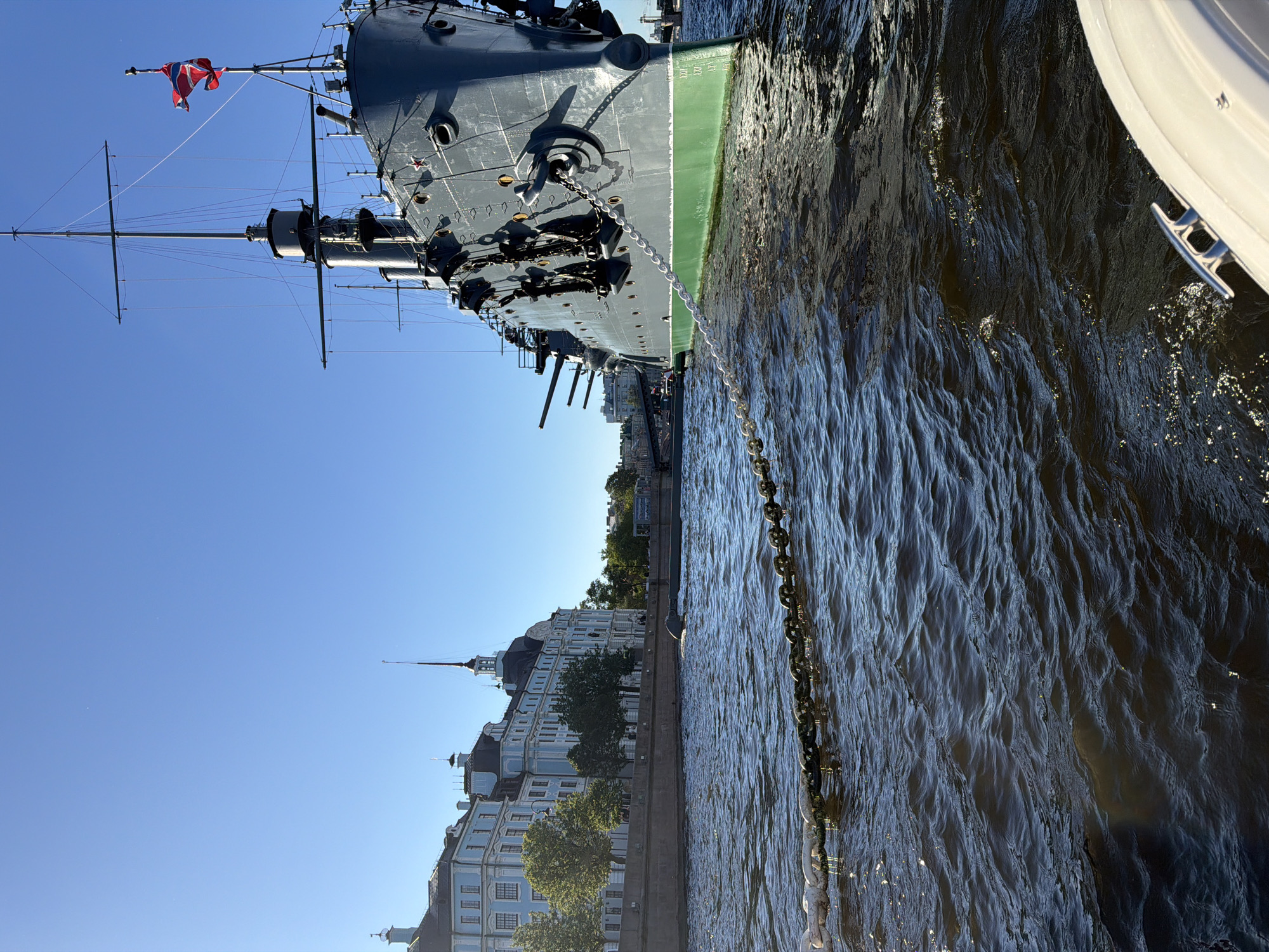 The image shows a large, grey warship docked in a body of water, likely a river or canal, in Saint Petersburg, Russia. The ship's mast and flag are visible, with a red, white, and blue flag flying. The ship's hull is painted a dark grey, with a distinct green stripe along the waterline. A heavy anchor chain stretches from the ship into the water, with a white boat or pontoon visible in the foreground on the right.
In the background, a row of ornate, light blue buildings with dark roofs lines the waterfront. These buildings appear to be historical and grand, characteristic of Saint Petersburg architecture. Trees are interspersed along the embankment, suggesting a park or promenade. The sky is a clear, bright blue, indicating a sunny day. The water is choppy, with sunlight glinting off the ripples. The perspective is from a slightly elevated position, looking down and across the scene. The image is oriented vertically, with the ship's mast pointing upwards and the buildings running horizontally along the bottom. The overall mood is peaceful and picturesque, capturing a moment of calm in a historic urban setting. No people are clearly visible in the image. No visible text can be deciphered.
