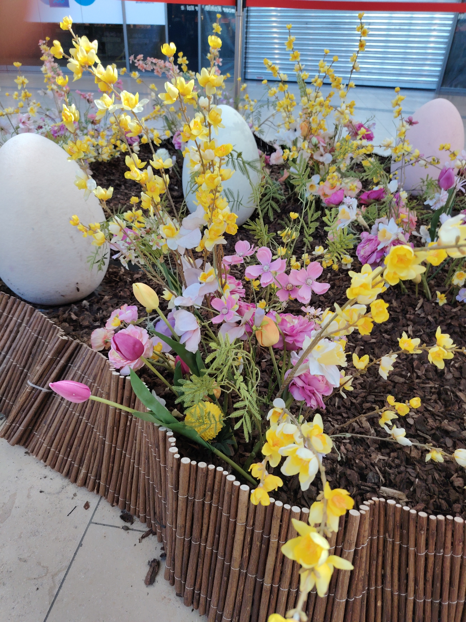 This is a close-up shot of an Easter display, likely located in the city of Kehl, Germany. The display features a circular flowerbed defined by a low fence made of thin, vertical bamboo-like sticks held together by string. Inside the flowerbed, there is dark mulch-like material.
The display includes a variety of artificial flowers, including pink and white blossoms, yellow flowers on branches, and tulips in various colors like pink, yellow, and peach. Three large, egg-shaped objects are placed among the flowers. Two are white, and one is pink.
The background is partially obscured, but a storefront with a horizontally ribbed or corrugated metal surface can be seen. There is also a sign in the upper part of the image with a red stripe across it. The pavement is visible in the foreground.