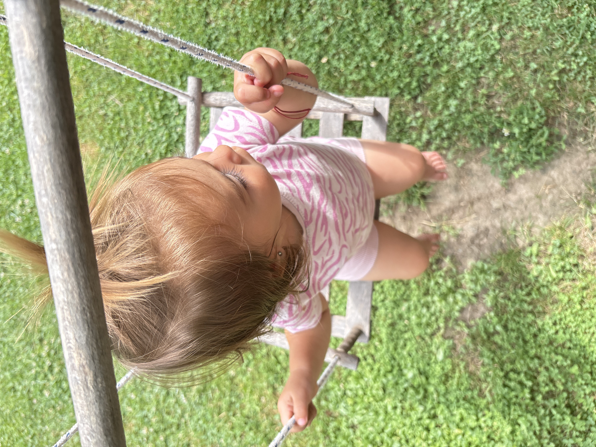 A young child with blonde hair tied in a ponytail is sitting on a wooden swing. The child is wearing a pink and white striped t-shirt and pink shorts. The child's hands are gripping the ropes of the swing. The swing is set in a grassy area. The image was taken from above the child. The location is Oreshak, Bulgaria.