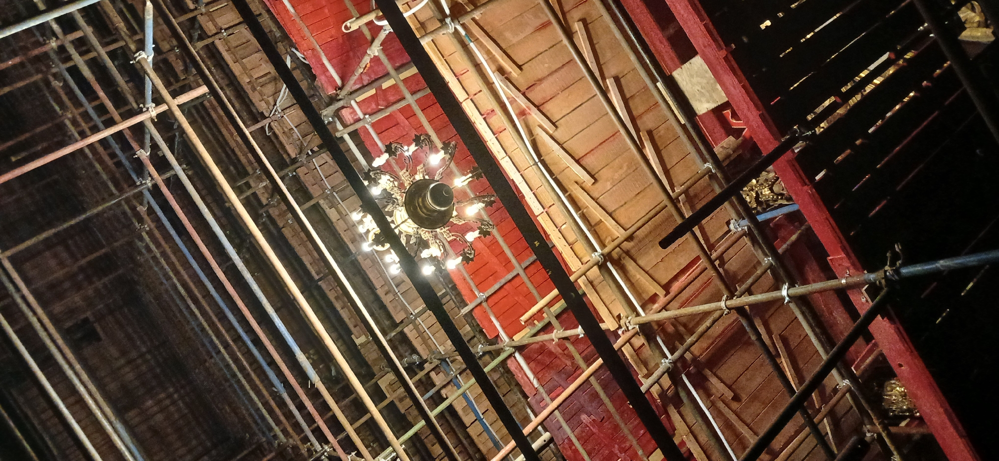 The image, taken from a low-angle perspective, depicts the interior of a building undergoing renovation, likely in Moscow, Russia. Visible is a complex network of metal scaffolding that dominates the foreground, crisscrossing the view toward the ceiling. In the center of the frame hangs an ornate, multi-tiered chandelier with numerous light bulbs, partially obscured by the scaffolding. The ceiling features wooden planks in varying shades of brown and red, some painted and some appearing unfinished. The environment appears to be dimly lit, emphasizing the scaffolding and the chandelier. No people are directly visible in the frame. The overall scene conveys a sense of construction and restoration within a historical building.