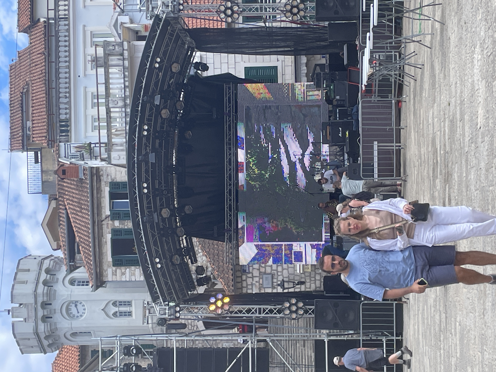 The image shows an outdoor concert stage set up in Herceg Novi, Montenegro. The stage features a large screen displaying colorful visuals, along with lighting and sound equipment. In the foreground, a man in a blue shirt and sunglasses stands beside a woman in white. Other people are milling around in the background. The backdrop includes traditional buildings with red-tiled roofs, characteristic of the Montenegrin architecture. A tower with a clock is visible to the left of the stage.