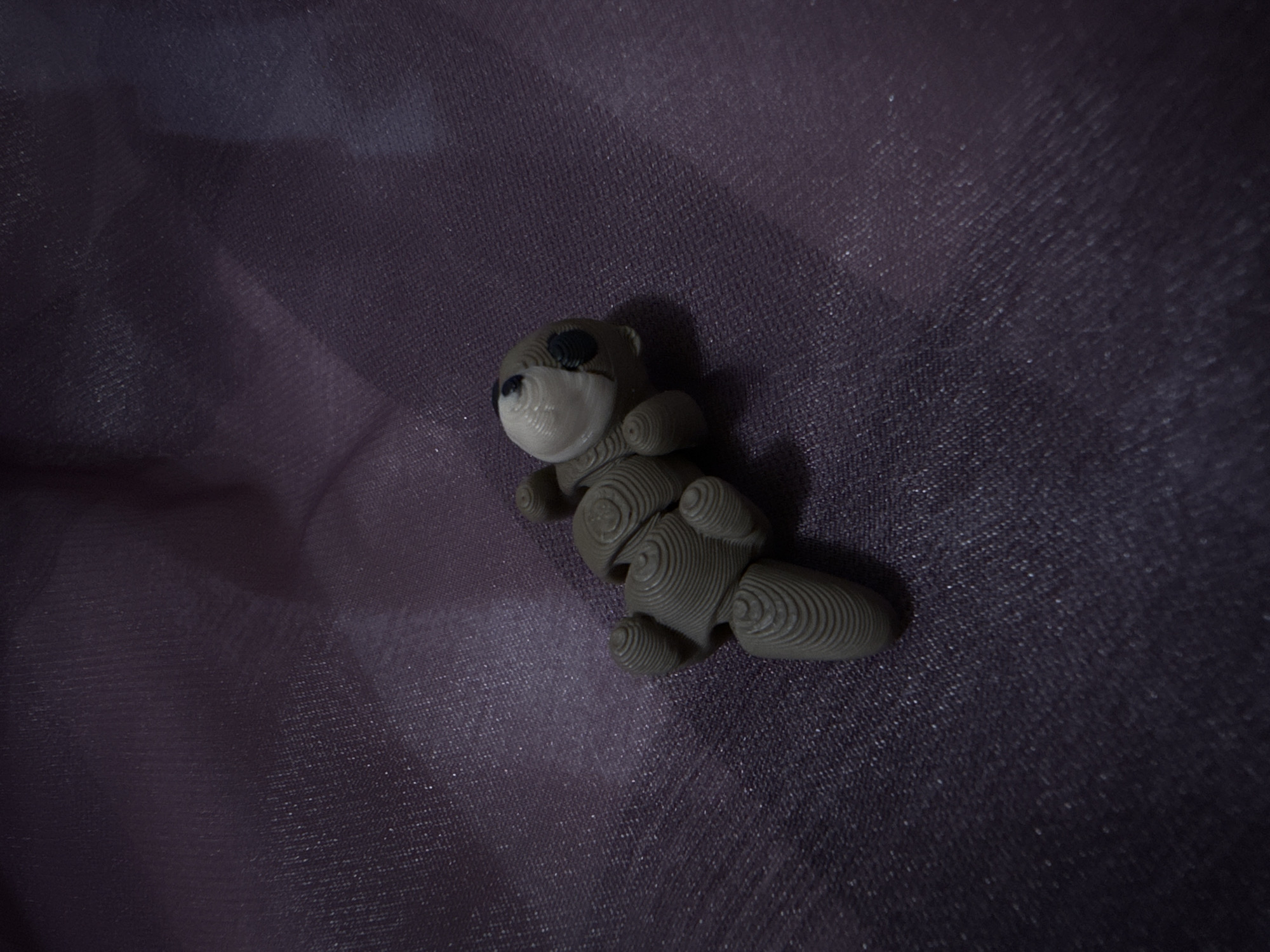 A 3D-printed toy bear sits on a dark-colored, textured cloth, likely polyester or a similar synthetic fabric. The bear is light gray and appears to be made from concentric circles of extruded material. It is lying on its side with its face towards the left. The lighting is uneven, with a brighter area on the left and darker areas on the right. The background cloth shows signs of being creased or wrinkled. The image does not provide location information about Puebla, Mexico.