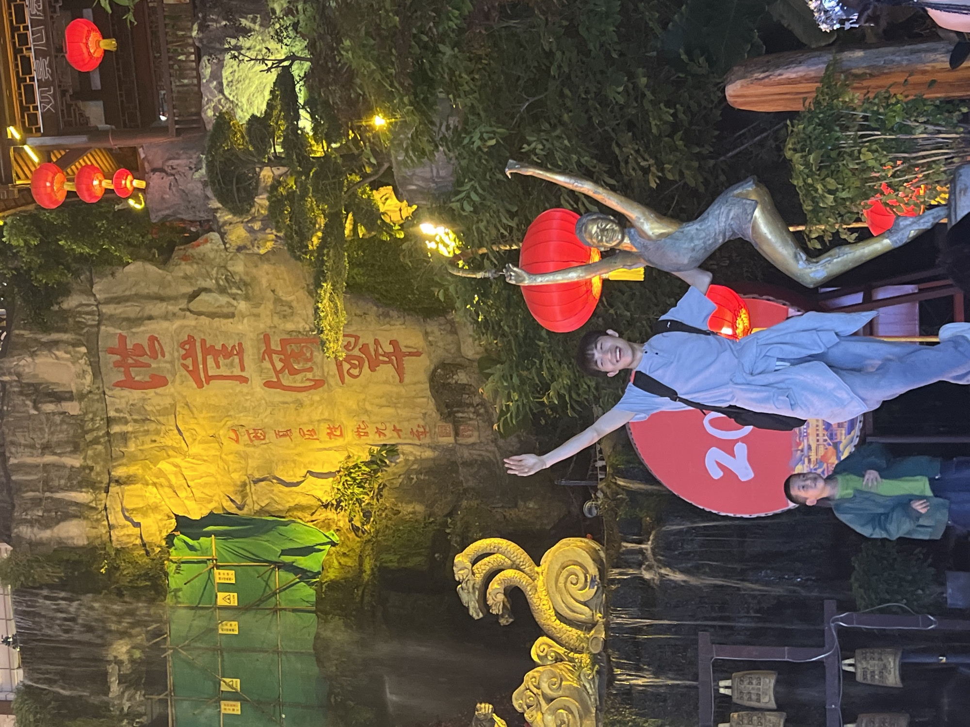 A nighttime outdoor scene shows a man standing with arms outstretched in front of a display in Yuzhong District, China. To his upper right is a bronze-colored statue of a figure seemingly in mid-flight, intersecting with a red lantern. The man wears a light blue outfit and a dark bag hangs across his torso. To his lower right stands a child in a green shirt and jacket. Behind them is a large red circle with the number "20" in white. Behind this, water is seen flowing. To the left is a rock formation, partly covered with green scaffolding. Chinese characters are prominently displayed on the rock face, illuminated by yellow light. Above the formation hang rows of red lanterns. Bells hang on a stand to the lower right. The scene appears to be a park or a tourist location.