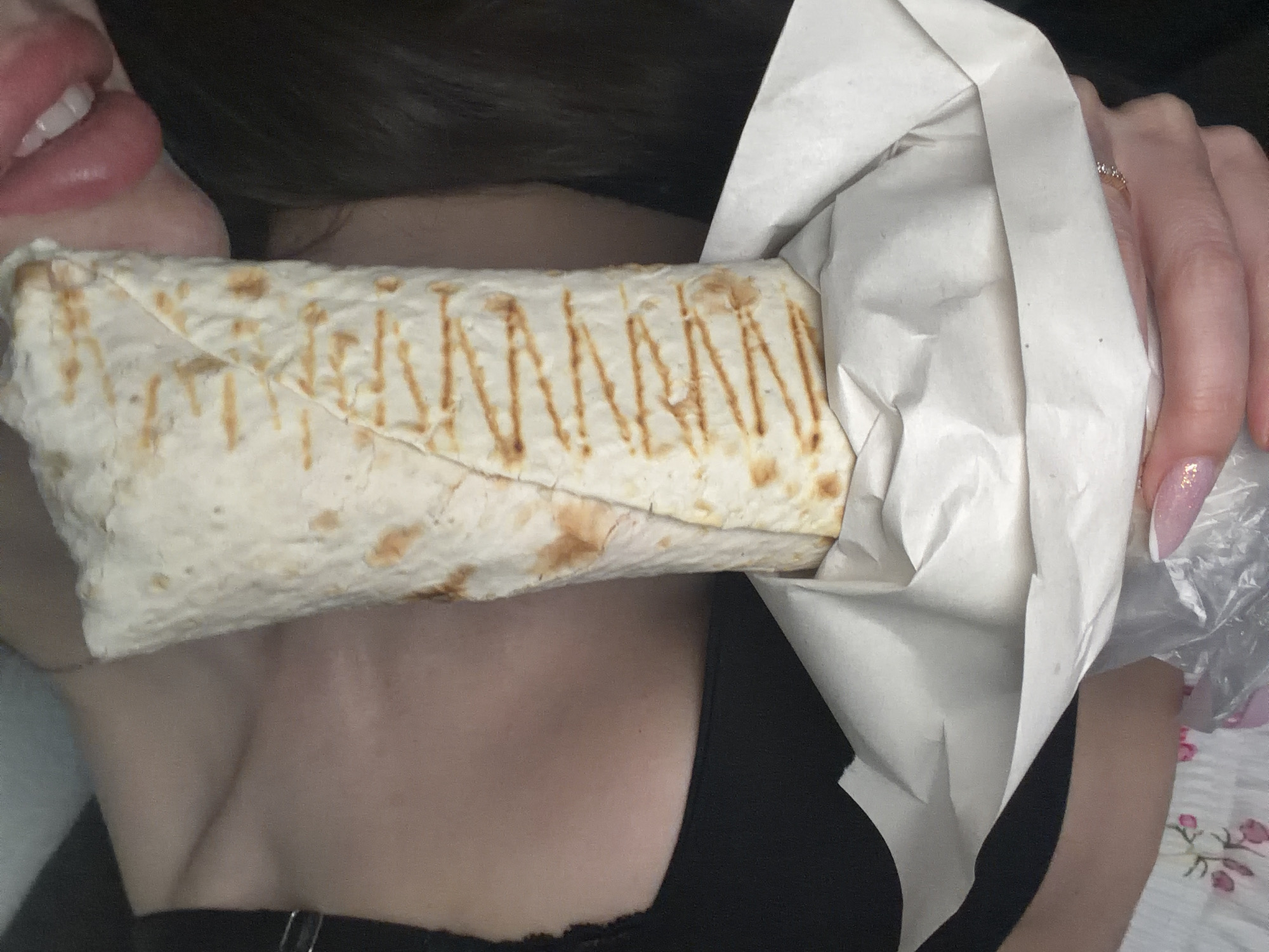 A close-up shot captures a fair-skinned person holding a shawarma wrap partially unwrapped in front of their face. The wrap is held in the left hand and is light in color, with darker grilled markings. The person's lips and teeth are visible, suggesting they may be about to eat the wrap. Their skin appears fair, and they wear a black top. A ring is visible on one of the fingers holding the wrap. The wrap is partially enclosed in white paper. The scene is indoors. No specific location in Saint Petersburg, Russia is discernible from the image.