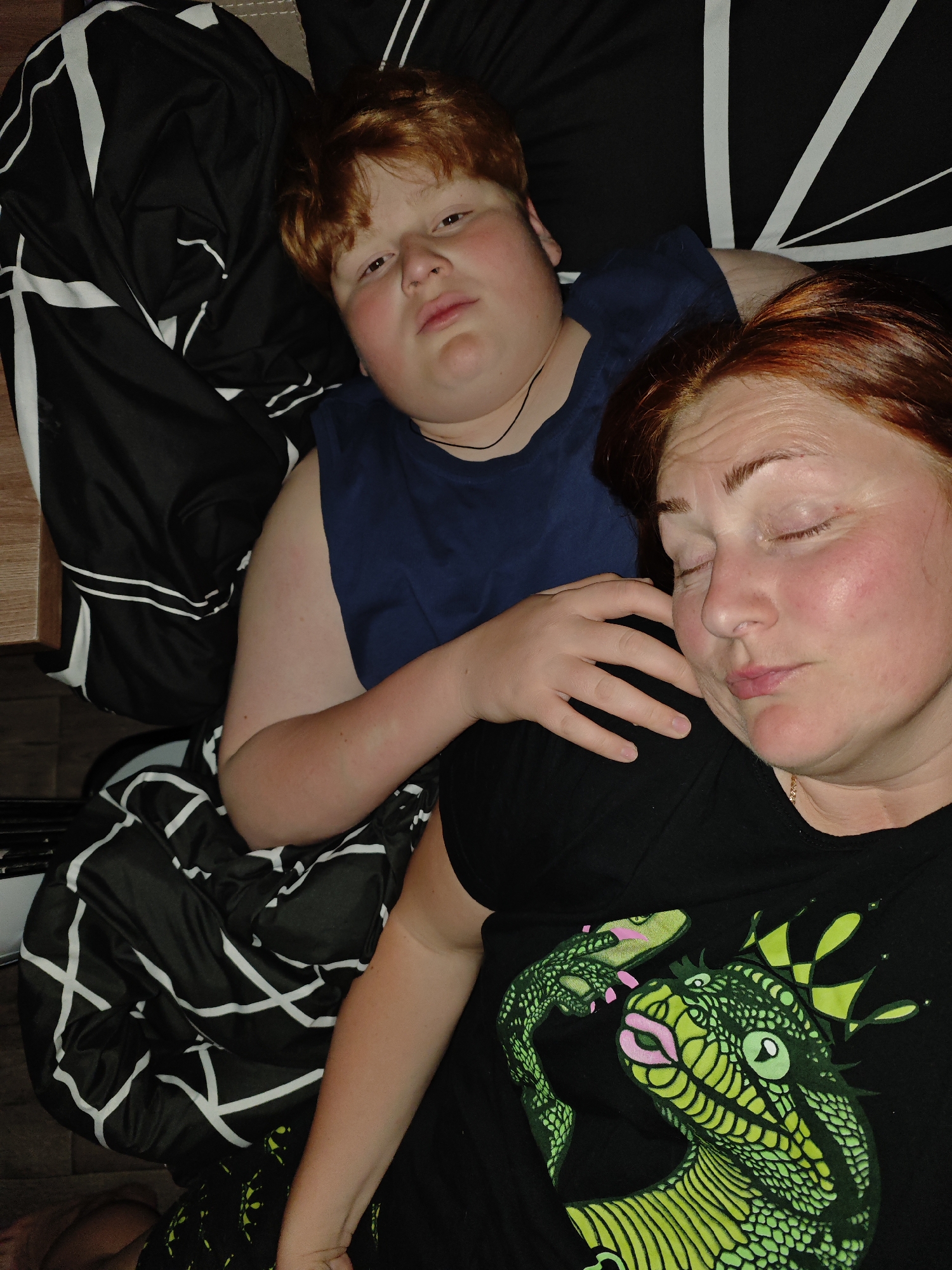 A woman with closed eyes lies next to a fair-skinned boy with reddish-brown hair, both on bedding of black and white. The boy is looking forward, wearing a blue shirt with his hand resting on the woman. The woman is wearing a black shirt with a green lizard design. A wooden nightstand is on the left of the image. The location of the image is not known.